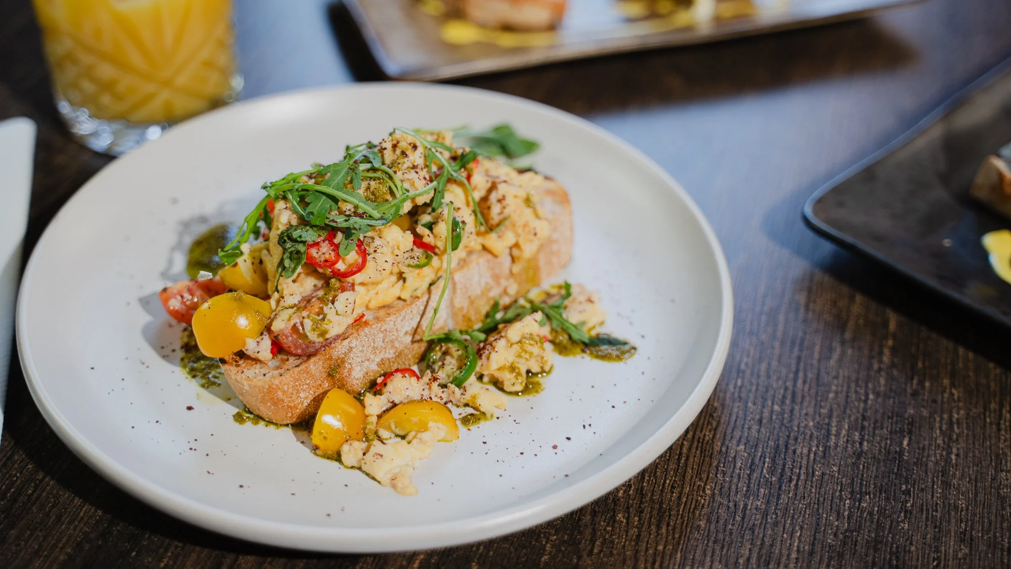A plate of bread topped with scrambled eggs, chopped tomatoes, red and green chili peppers, fresh arugula, and drizzled with green sauce, set on a dark wooden table.
