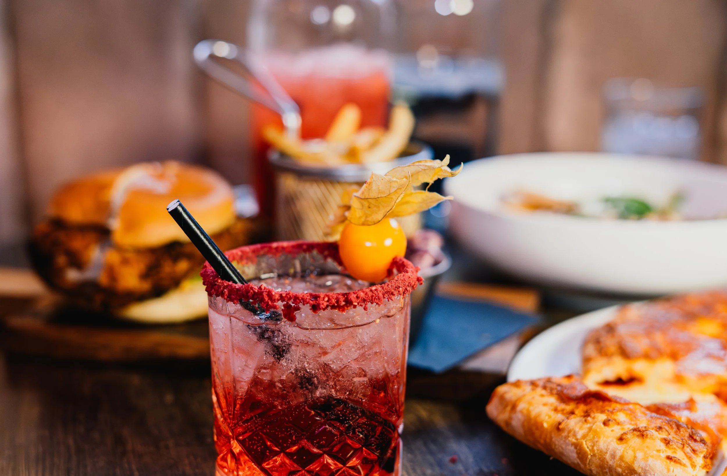 A pink cocktail with a salted rim, garnished with a yellow cherry and a leaf, on a table with food including fries, a burger, and pizza in the background.