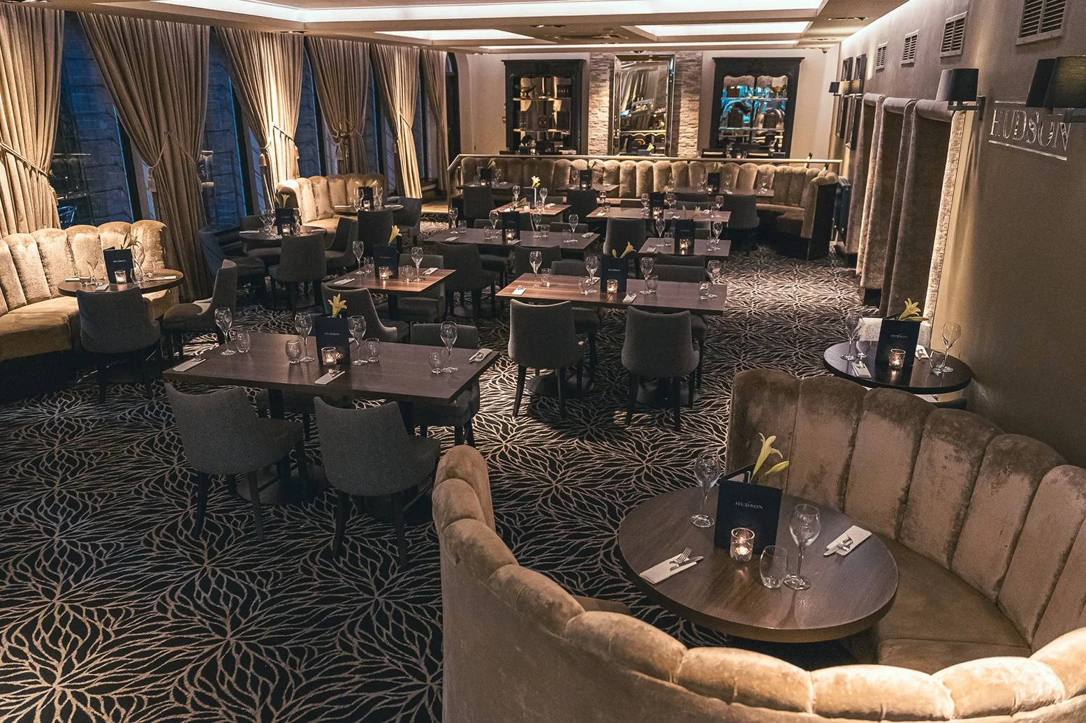 Elegant dining room with dark wood tables, upholstered chairs, and round booths, set with wine glasses, candles, and flowers. Large windows with gold curtains and a patterned carpet.