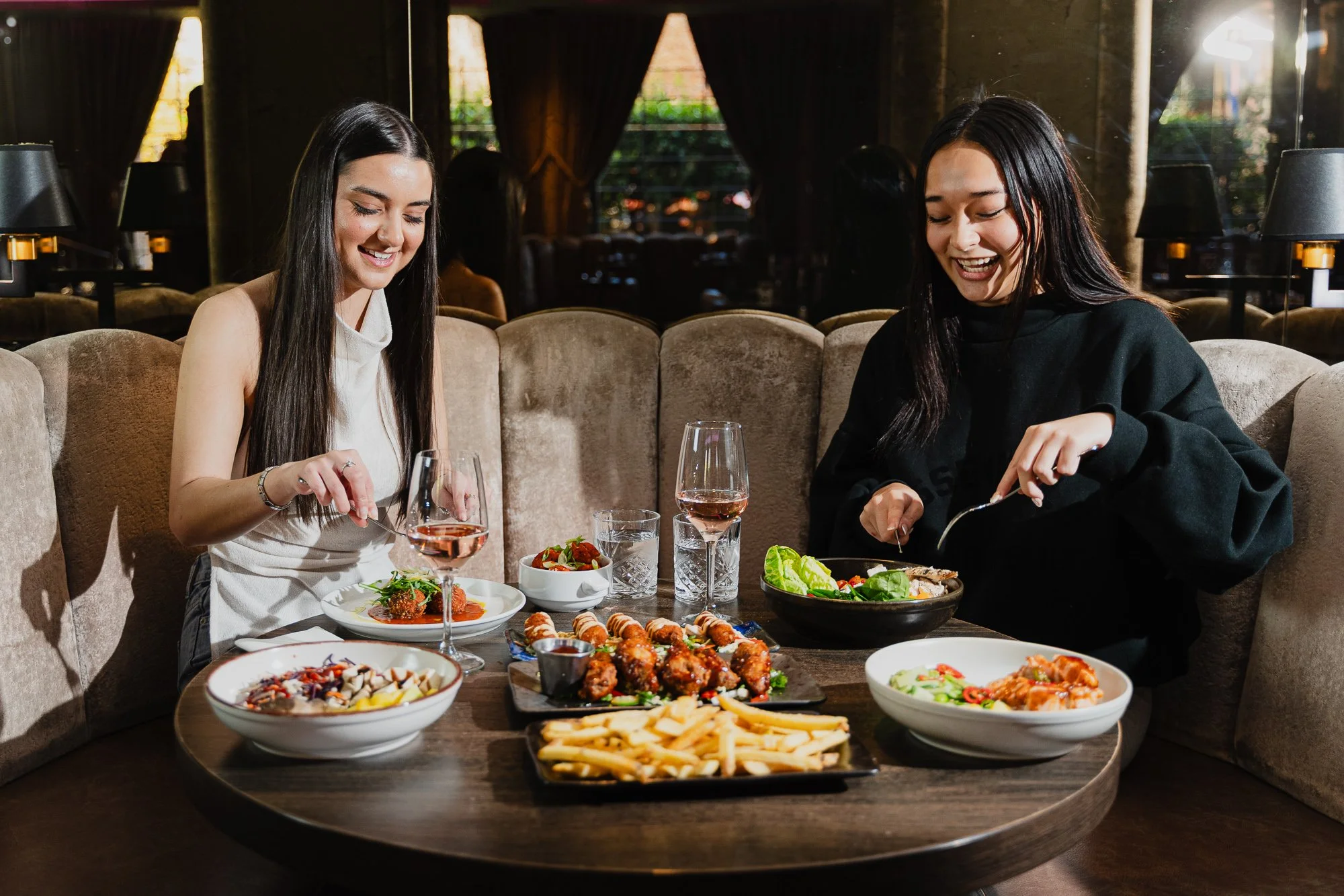 Two women are happily eating at a restaurant with a large spread of various dishes, including salads, fried foods, pizza, and drinks, on a round wooden table and beige cushioned seating.