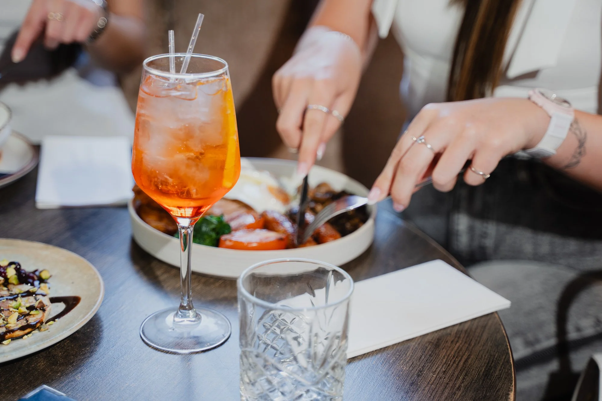 A tall glass of orange aperitif with ice and a straw on a dining table with various plates of food, including a pizza and a bowl of salad, in the background.