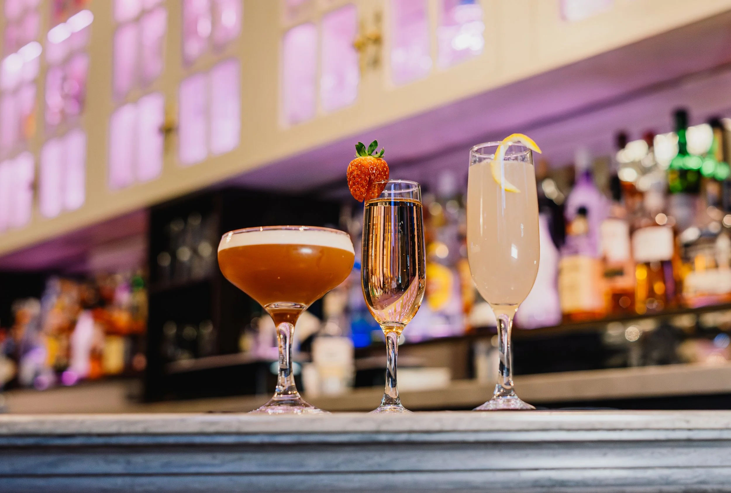 Four cocktails on a bar counter with a blurred bar background. The drinks include a maroon-colored cocktail with a straw, a champagne flute with lemon slices, a bubbly drink with a strawberry garnish, and a pinkish cocktail with an orange slice.