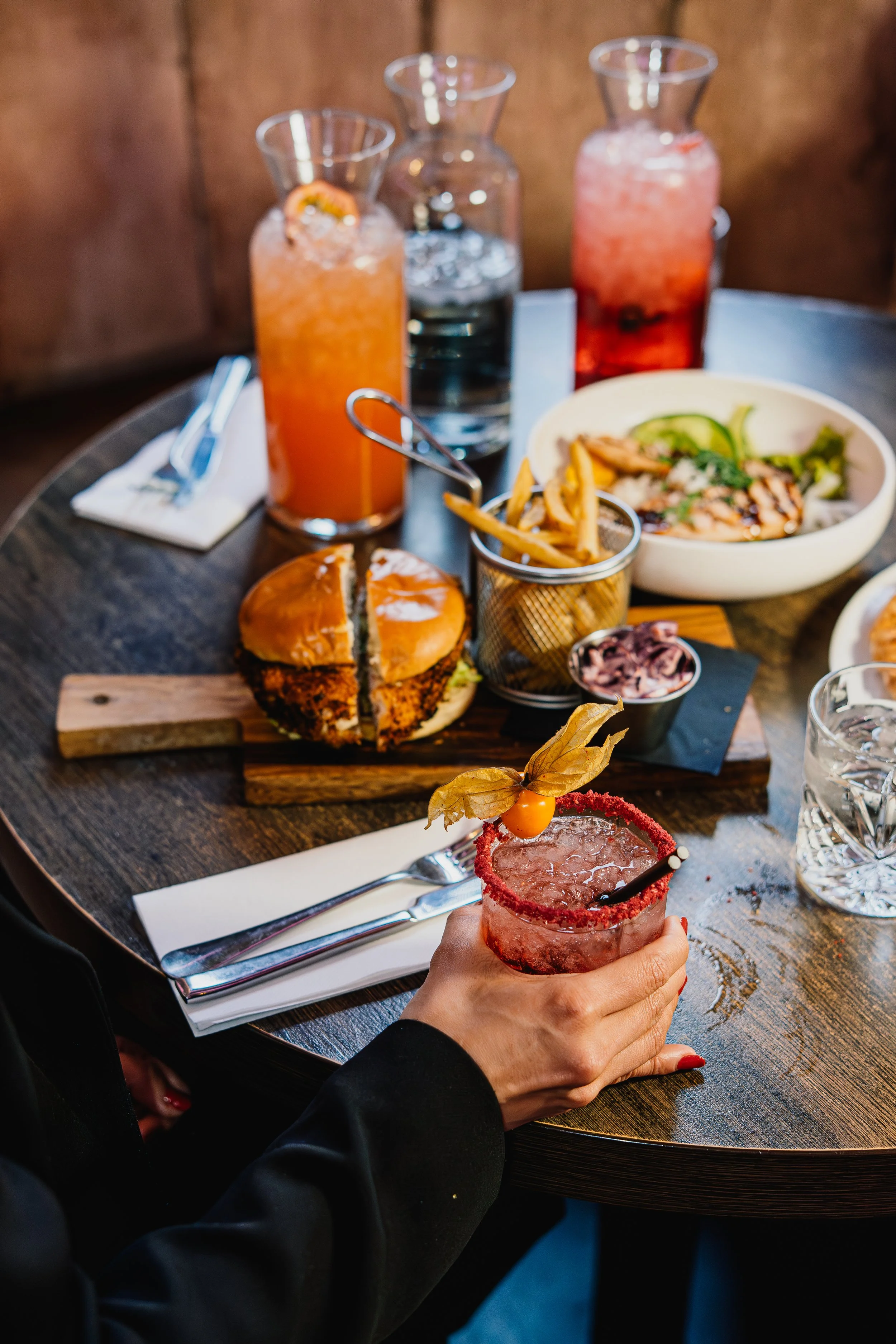 A person with red nail polish holding a pink cocktail garnished with a physalis fruit in a glass rimmed with red sugar. In the background, there is a table with three pitchers containing colorful drinks, a plate of food, and a small dish with crispy fries. The table is set with napkins and cutlery.