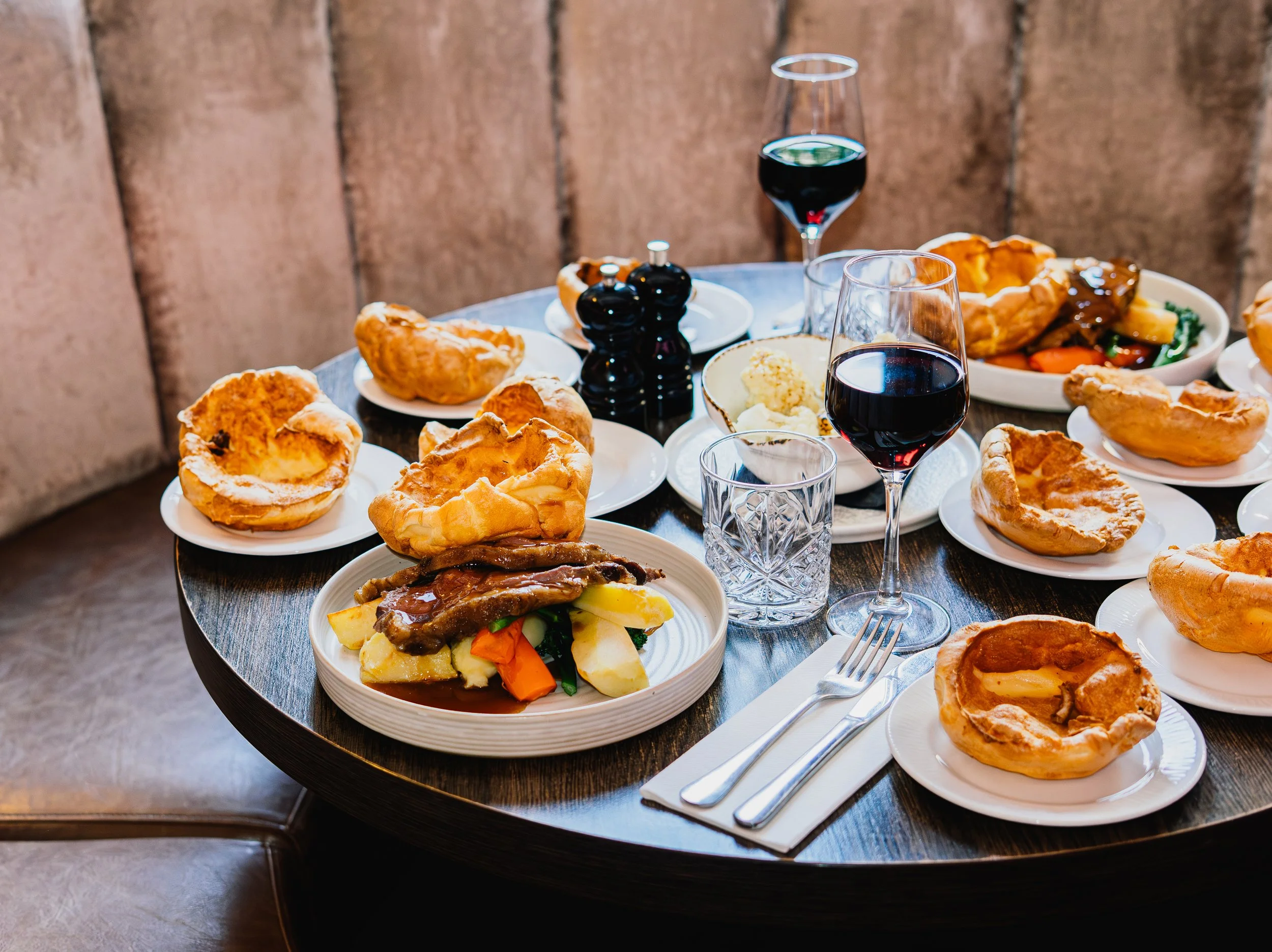 Table with various plates of food including Yorkshire pudding, roasted vegetables, a bowl of mixed vegetables, and glasses of red wine. The background features a wooden wall.