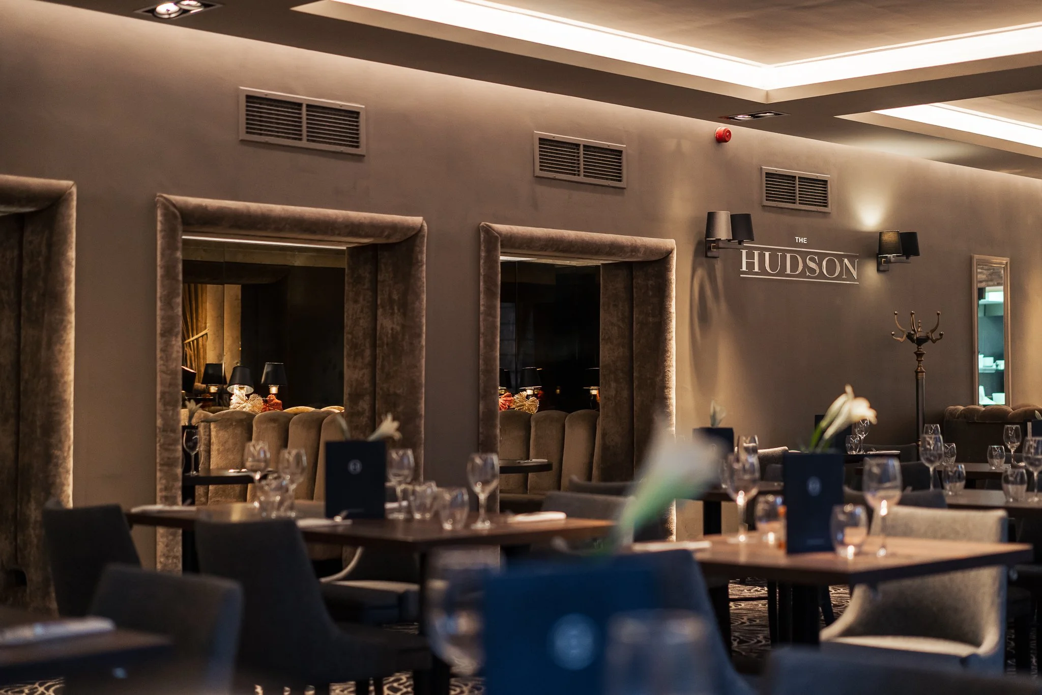 Interior of a restaurant or lounge with tables set for dining, decorative lighting, mirrors on the wall, and a sign that reads 'The Hudson'.