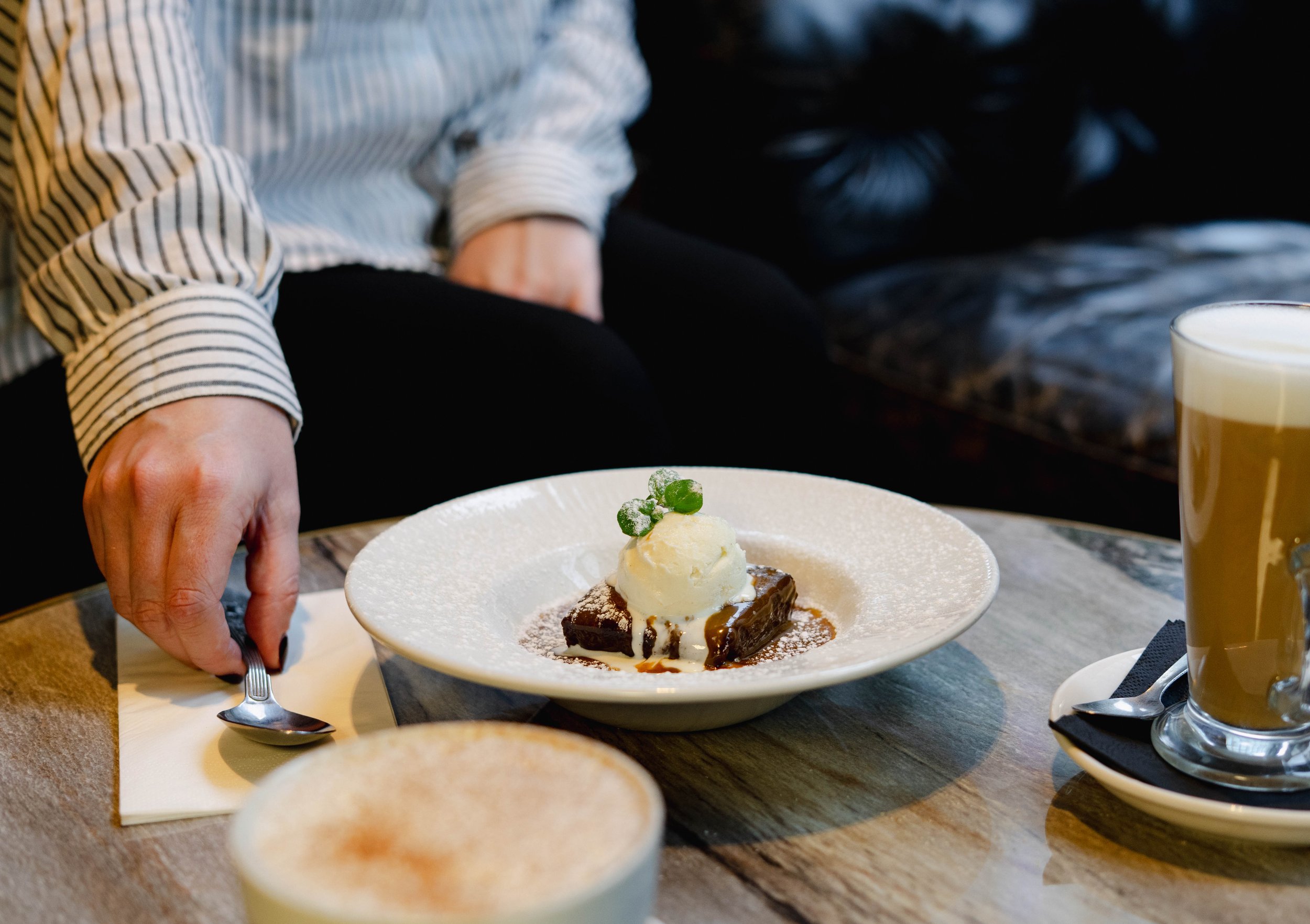 Person reaching with a spoon towards a dessert with ice cream, caramel, and mint on a plate. A glass of iced coffee is on the table.