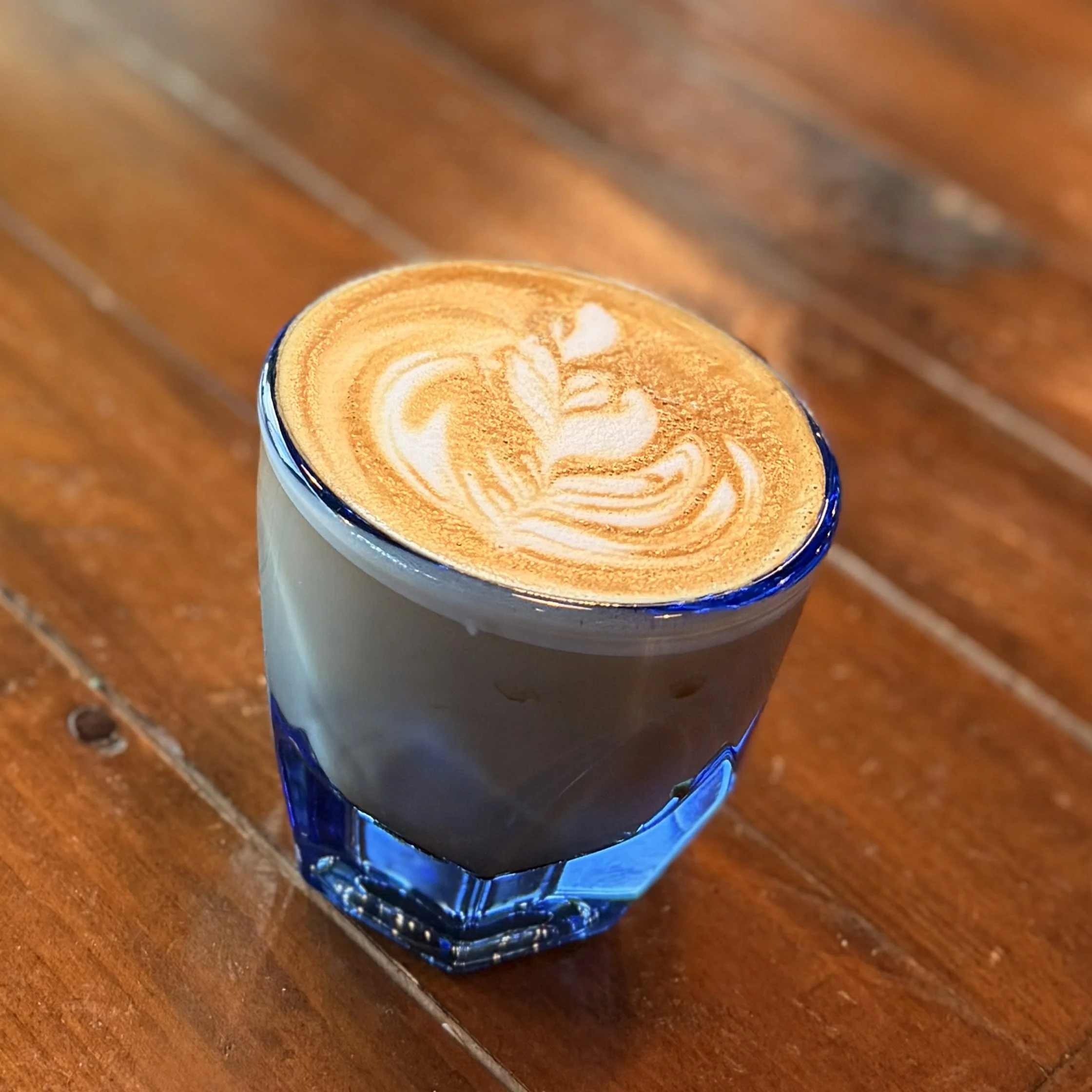 Close-up of a latte with latte art in a blue glass mug, sitting on a wooden table.