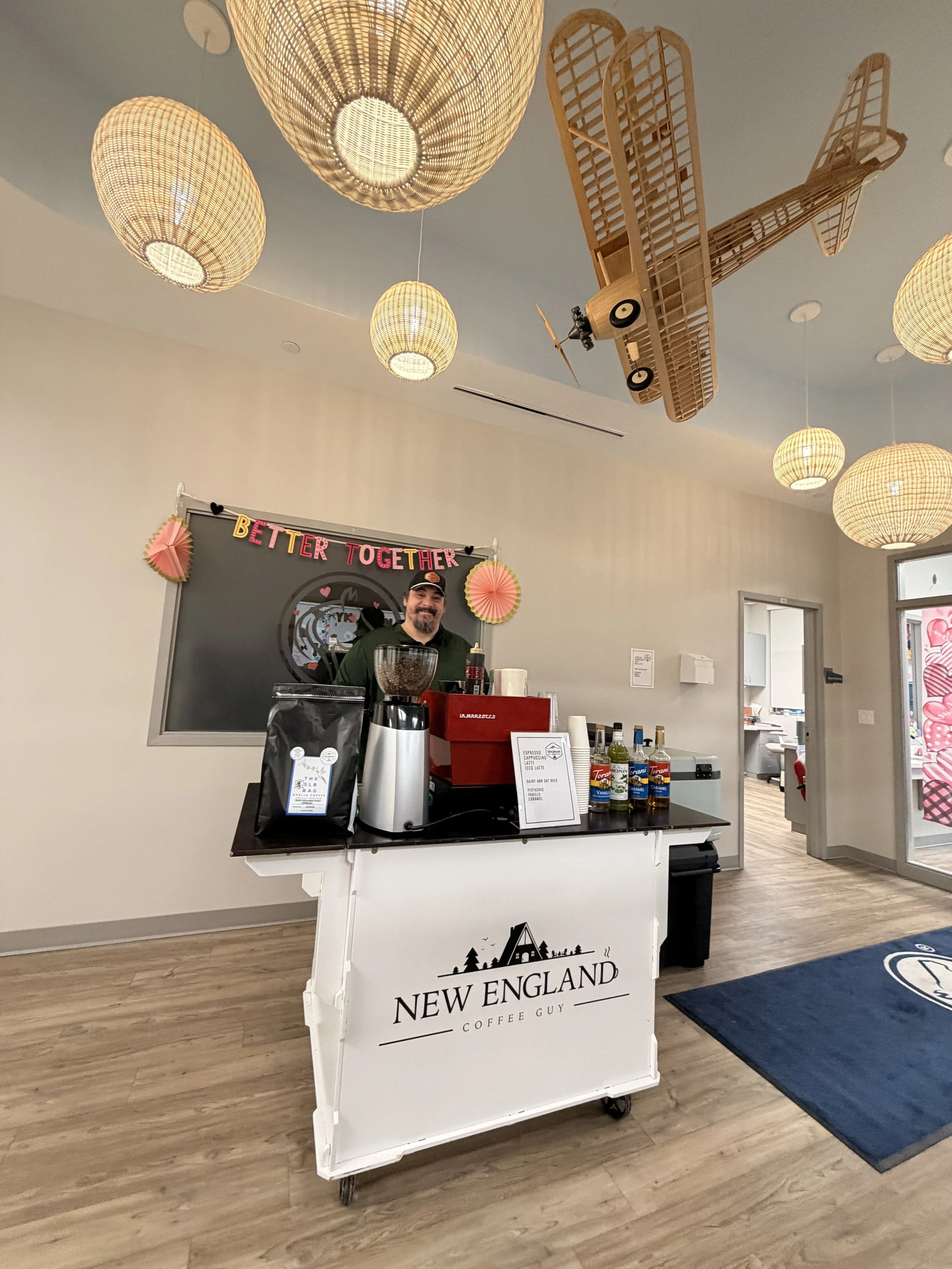 Coffee stand with a man behind it, displaying various bottles and coffee-making equipment. The stand has a sign that says 'New England Coffee Guy' and is in a room with hanging wicker lights and ceiling decor shaped like vintage airplanes.