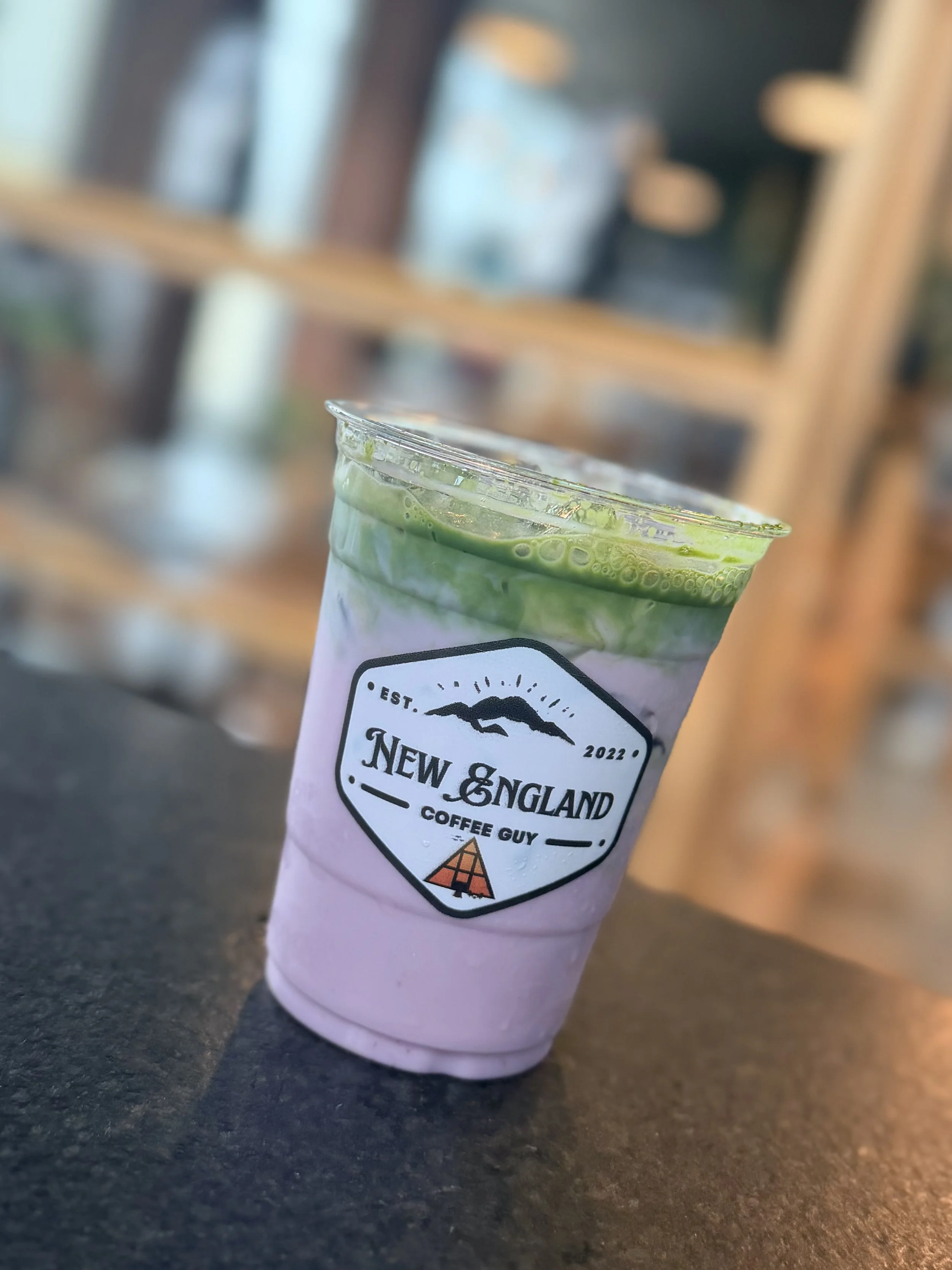 A pink paper cup with a logo that reads "New England Coffee Guy," containing a green iced drink with bubbles, placed on a dark surface with a blurred background.