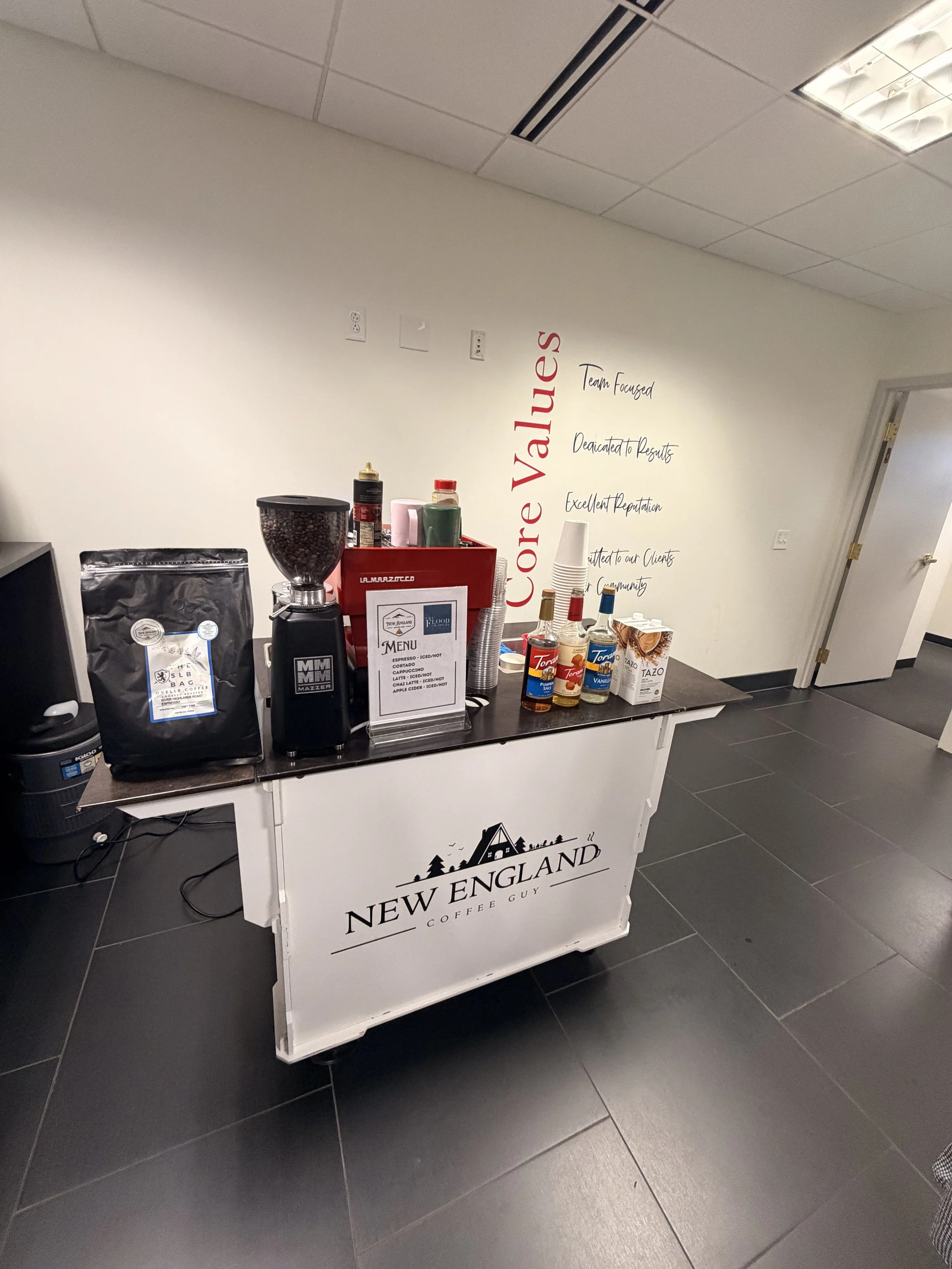 Coffee station with various coffee and condiment bottles, a coffee grinder, and a sign that reads "New England Coffee Guy." The station is set up against a plain white wall with a large red and black inspirational quote.
