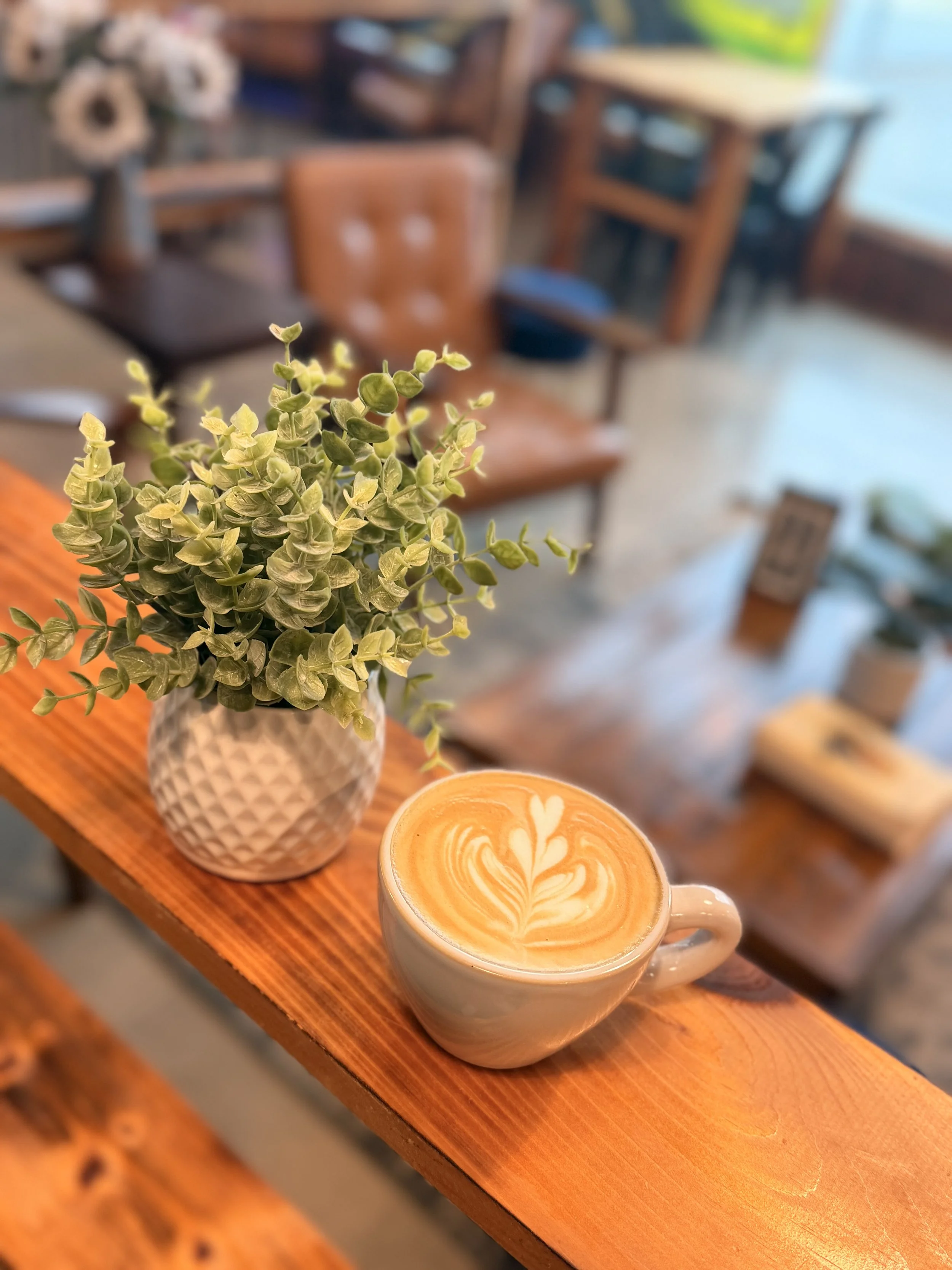 Close-up of a latte with latte art in a white mug on a wooden table, with a pot of flowers and a plant pot in the background inside a cozy cafe.