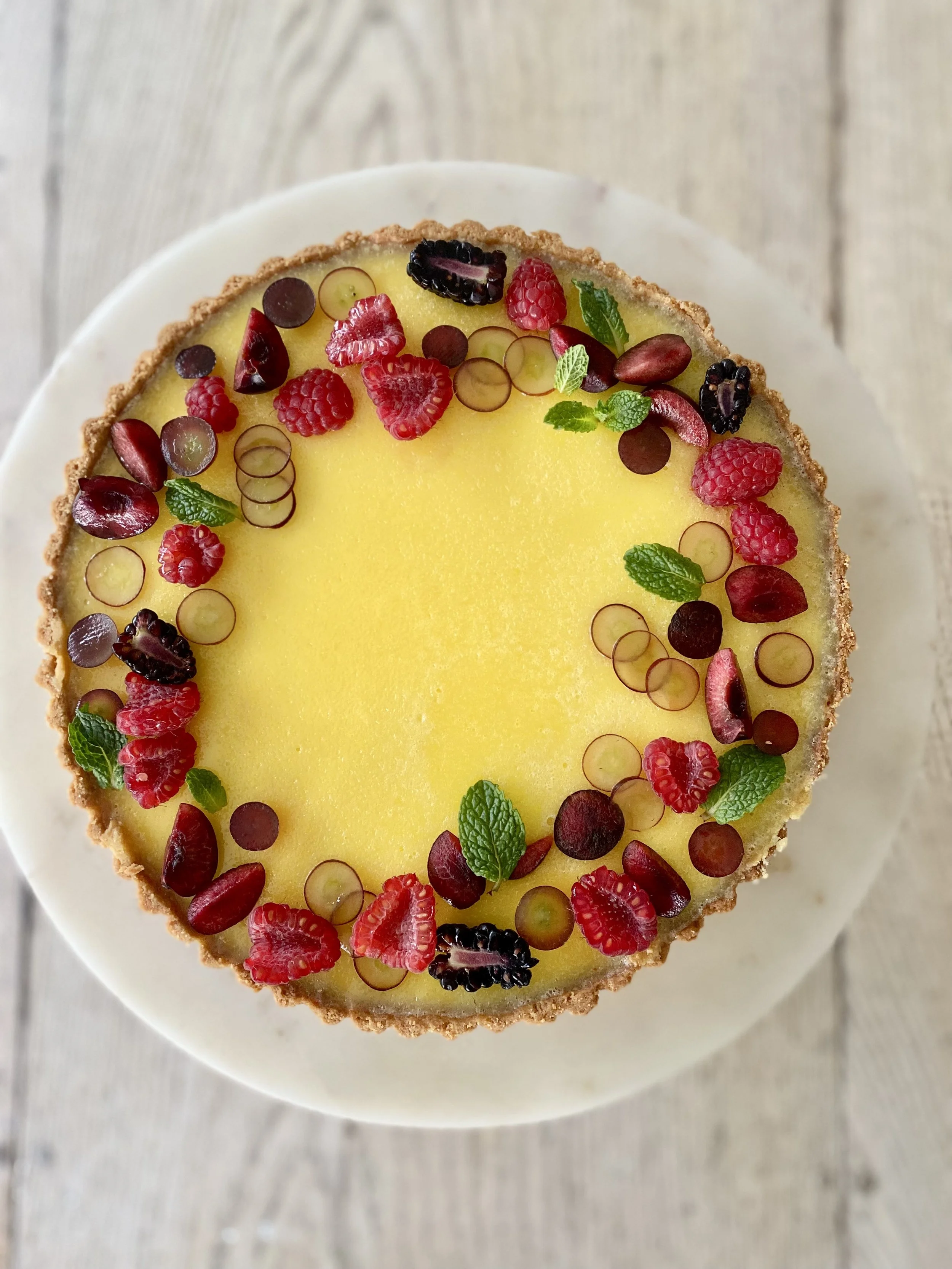 A top view of a round fruit tart with a yellow filling, decorated with various berries and mint leaves on a white plate, placed on a light wooden surface.