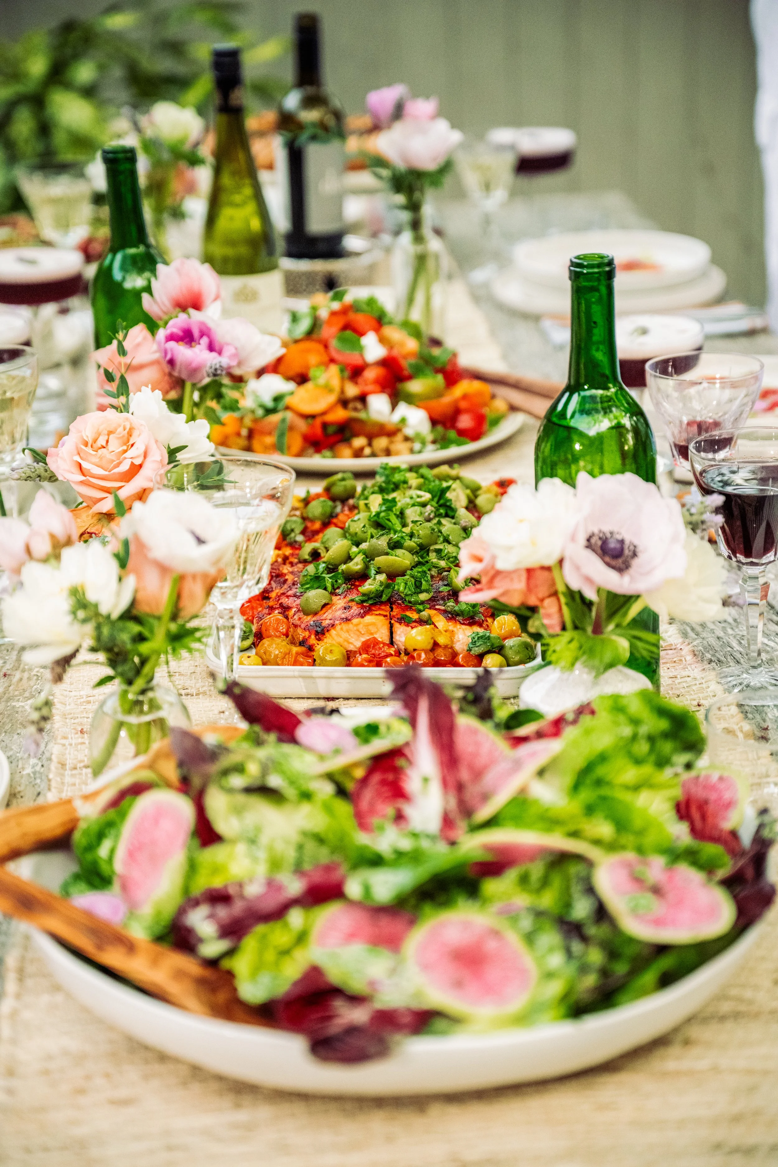 A colorful table setting with flowers, bottles, and a variety of dishes including salads and a platter with cooked vegetables and greens.