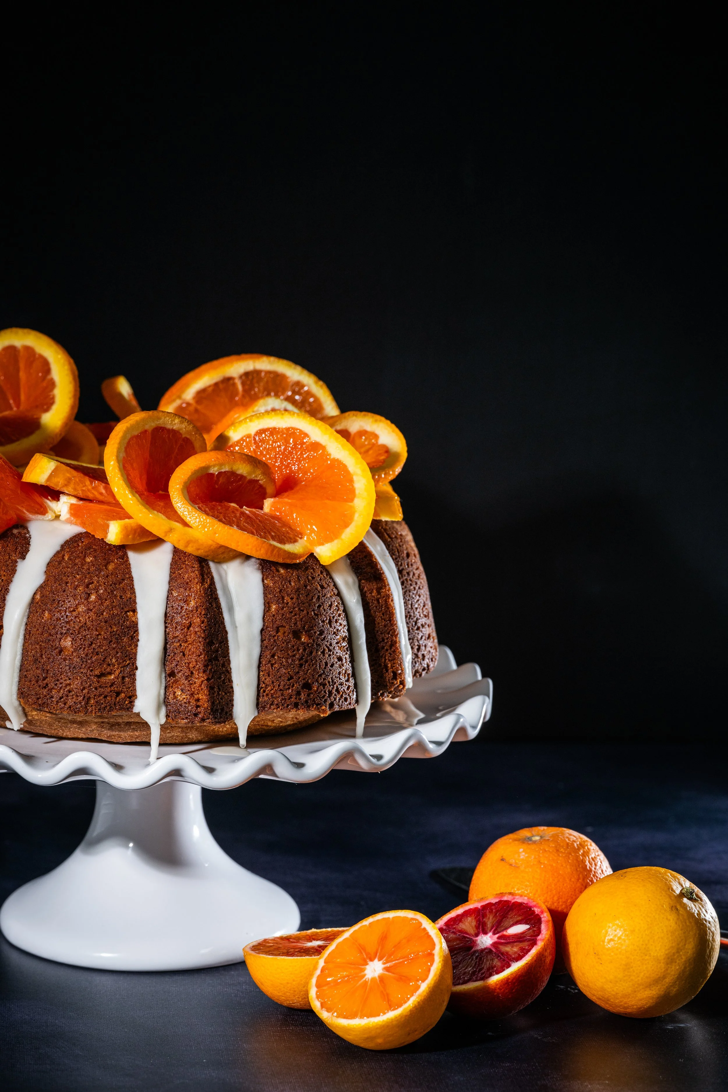 A bundt cake decorated with orange slices and drizzled with white icing, placed on a white cake stand against a black background, with additional oranges and blood oranges in front.