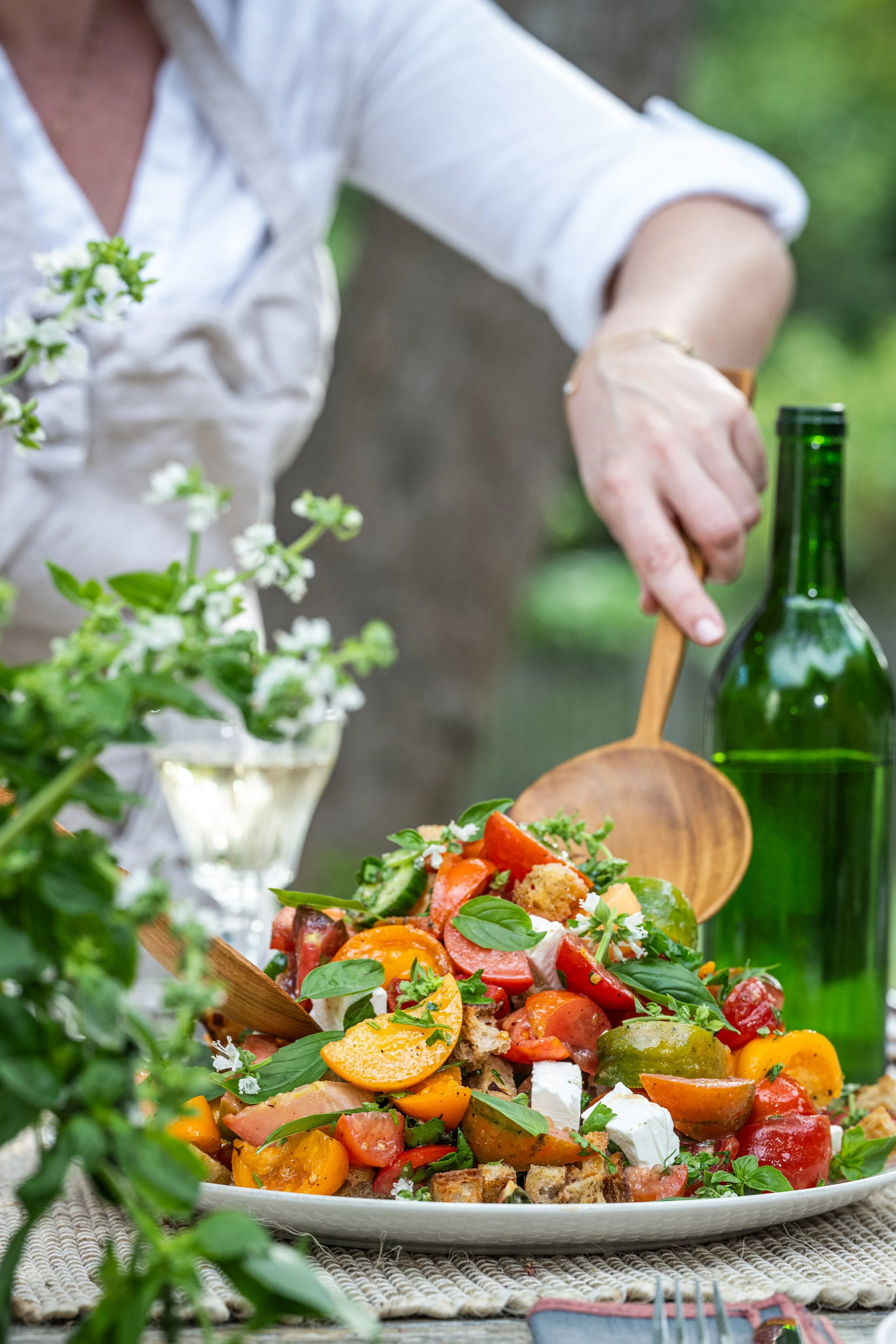 A person serving a colorful vegetable salad outdoors with a wooden scoop, surrounded by greenery and a green glass bottle.