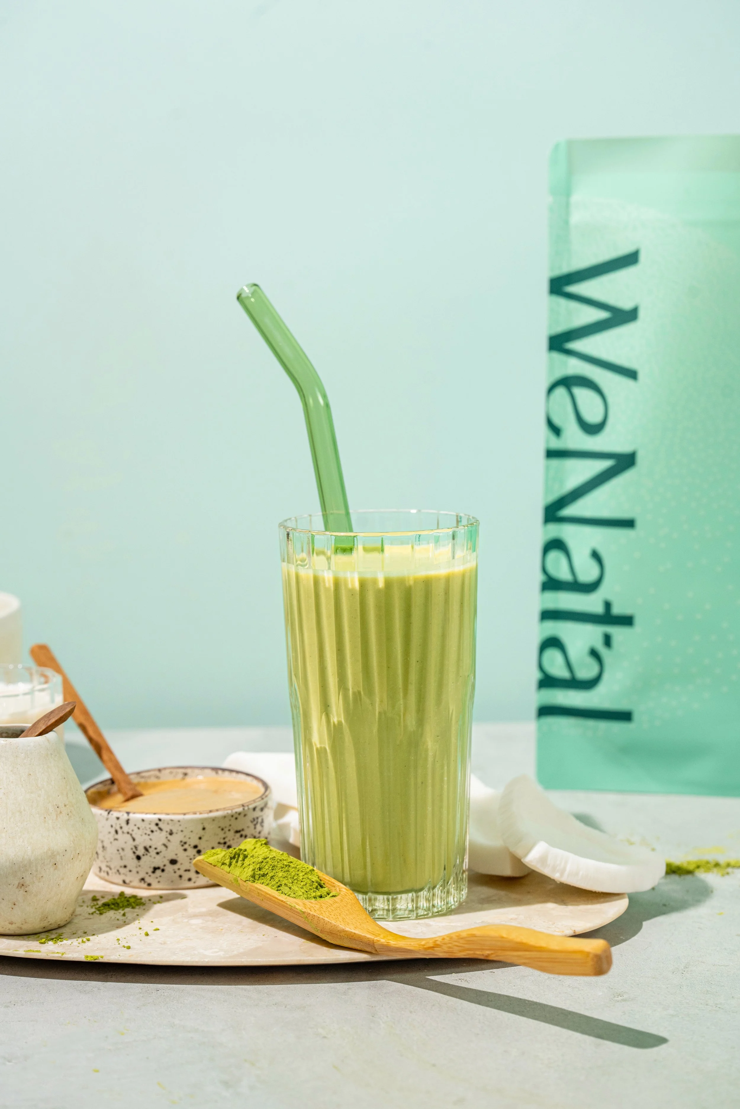 A tall glass of green smoothie with a green straw, surrounded by matcha powder, a small bowl with a spoon, and other matcha ingredients on a marble tray, with a mint green ValpaNale shopping bag in the background.