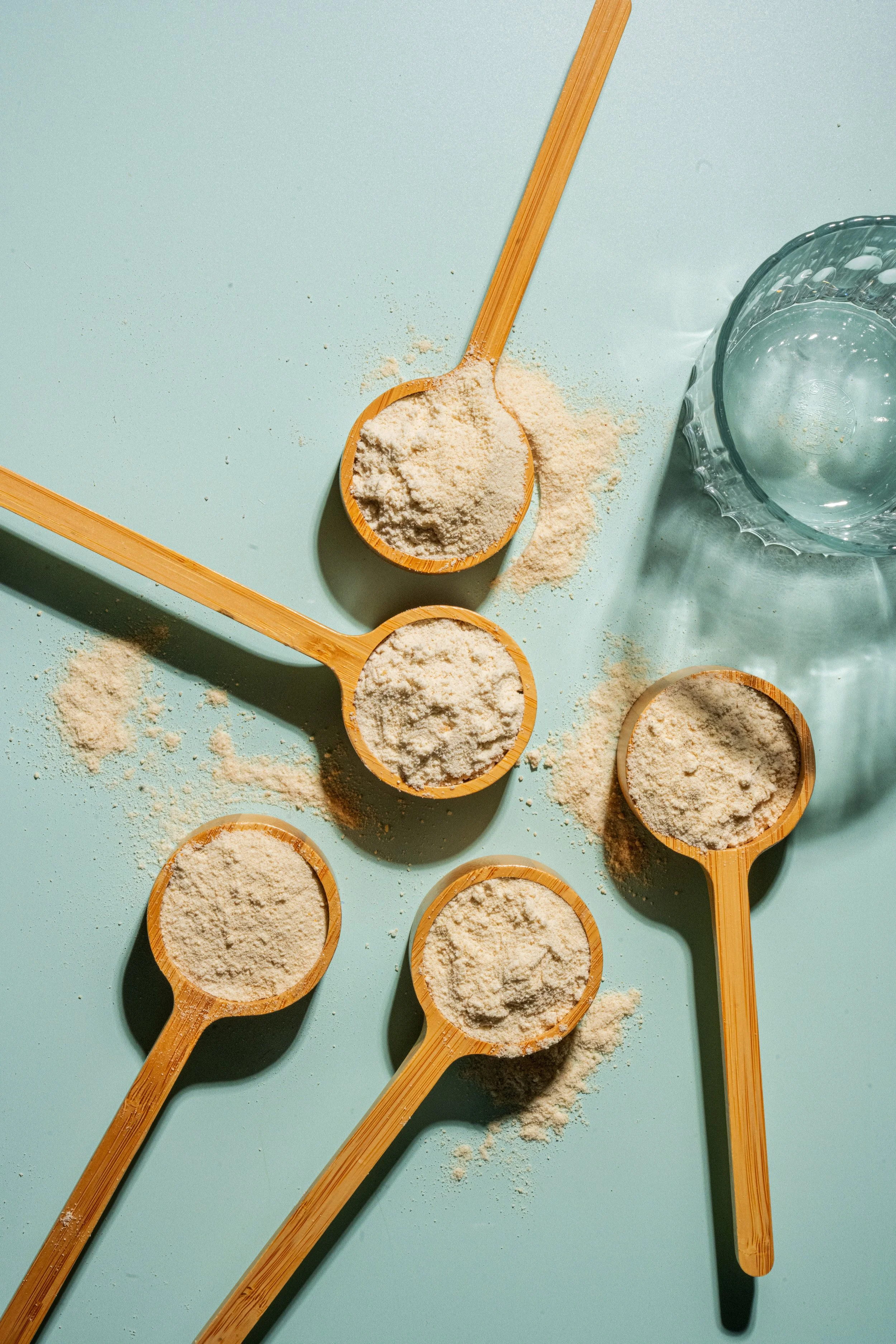 Multiple bamboo measuring spoons filled with flour scattered on a mint green surface, with a glass of water nearby.
