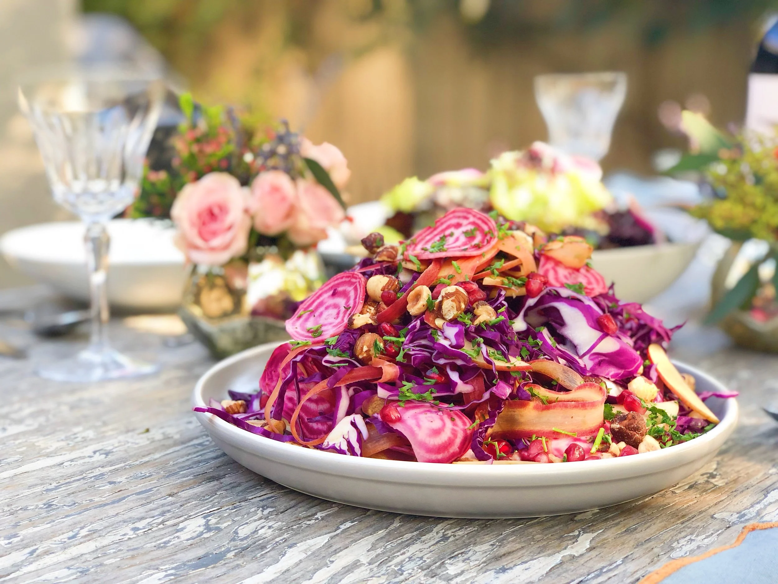 Colorful mixed vegetable salad with purple cabbage, radishes, carrots, nuts, and green herbs on a white plate, outdoors on a rustic wooden table, with flowers and glasses in the background.