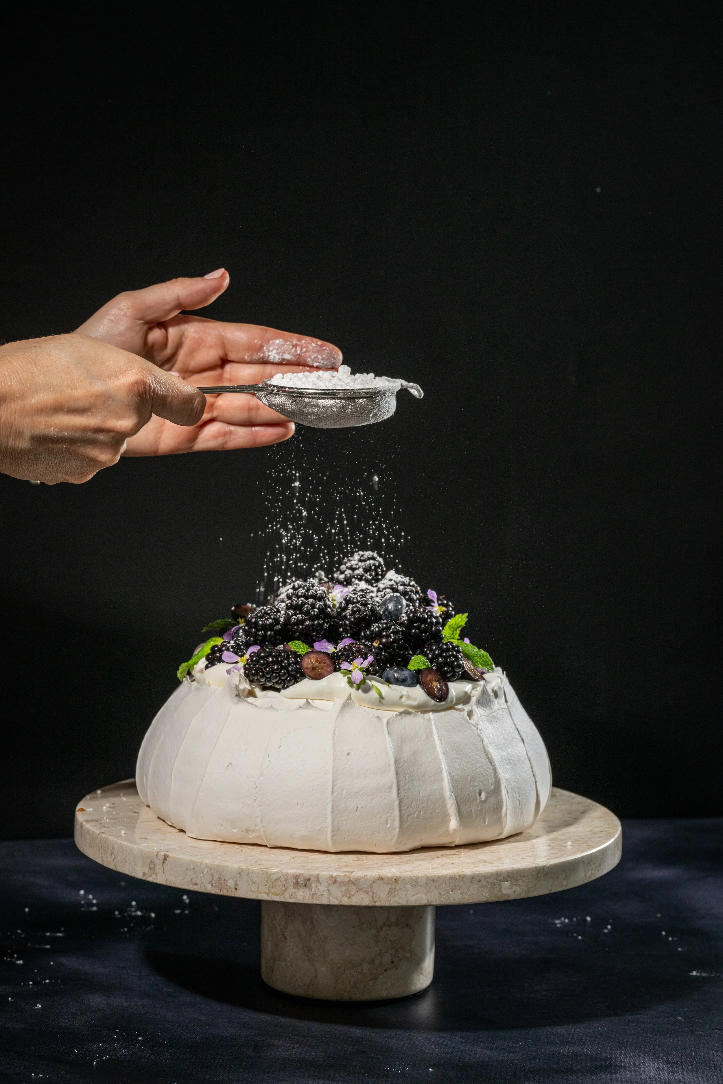 A baker sifting powdered sugar over a decorated pavlova topped with blackberries, blueberries, grapes, edible flowers, and mint leaves, on a marble cake stand against a black background.
