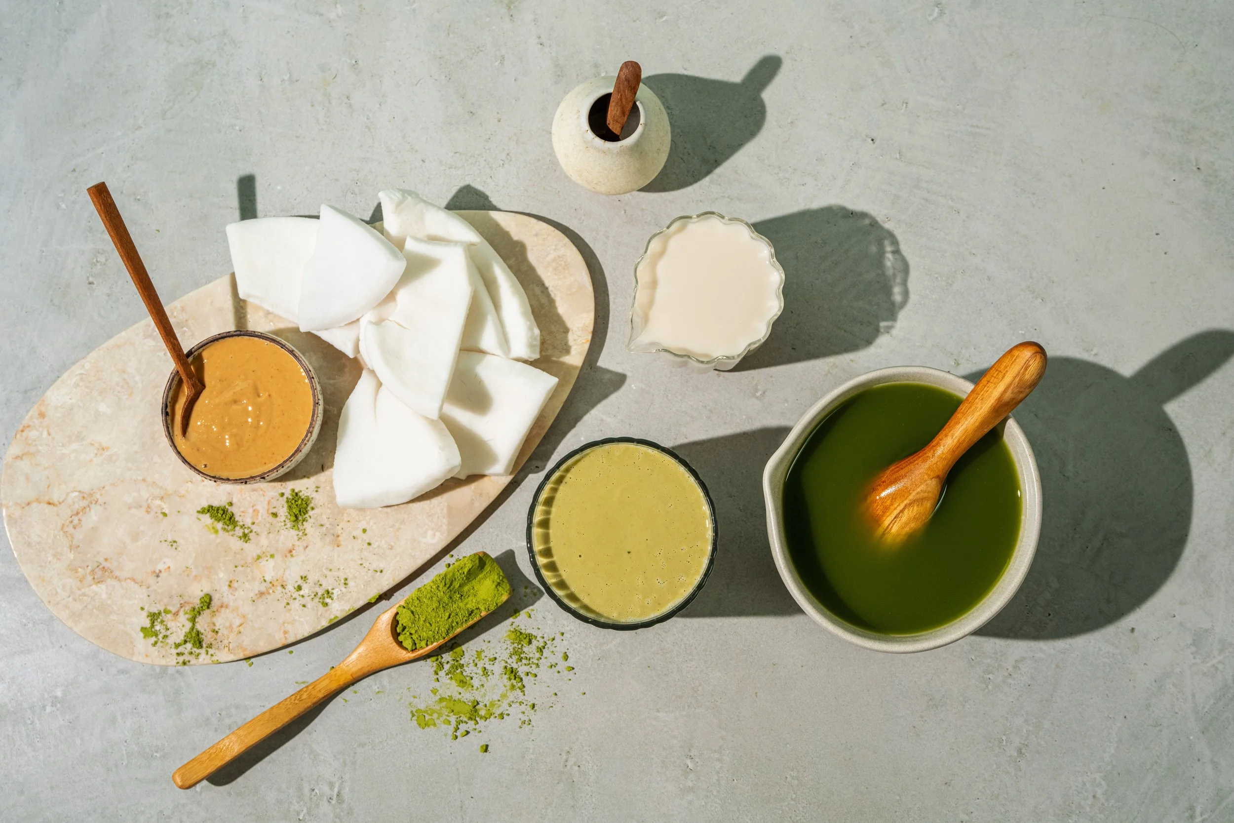 Assorted ingredients and liquids for preparing Matcha tea, including whipped coconut milk, ground green tea powder, a bowl of green tea, a bowl of matcha latte, a small container of sweetened condensed milk, and a rock salt grinder on a light surface