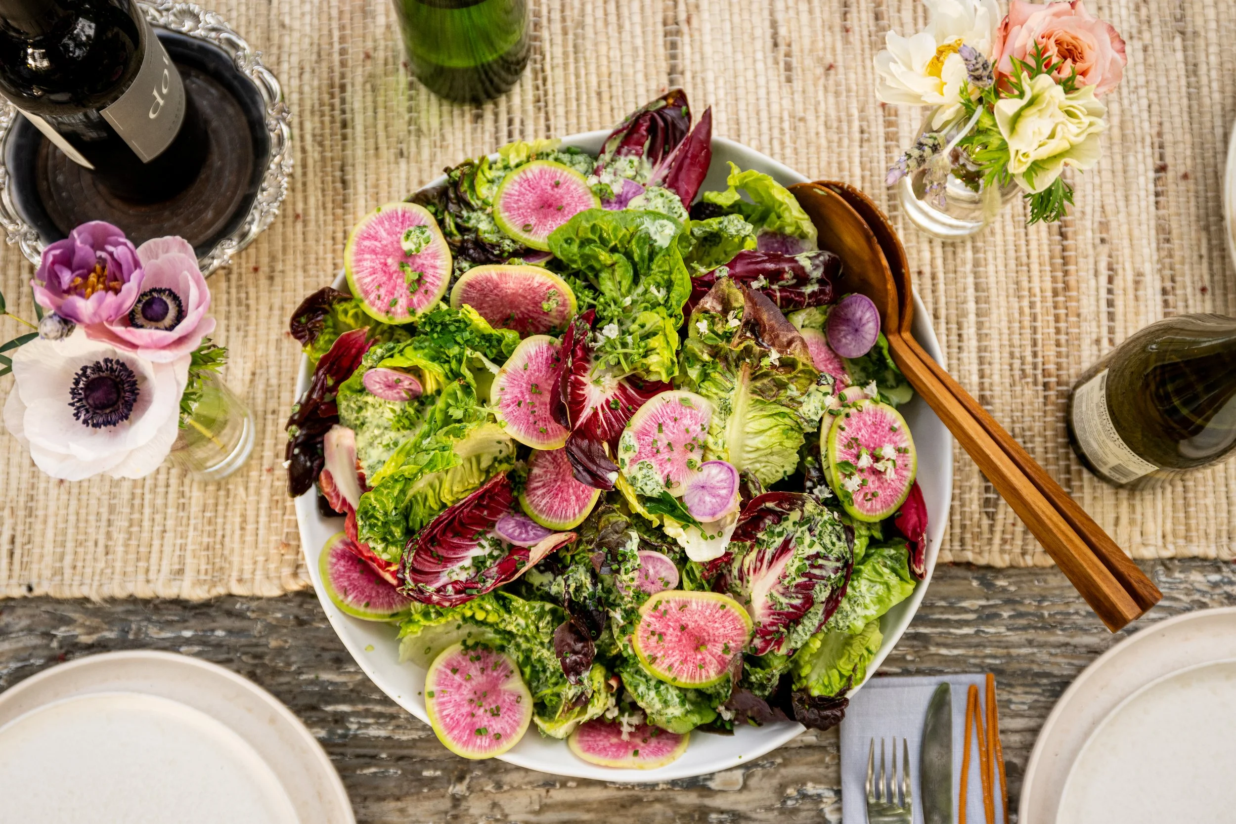 A large bowl of mixed greens salad with sliced watermelon radishes, served on a wooden table with wine bottles, glasses, flowers, and empty plates.