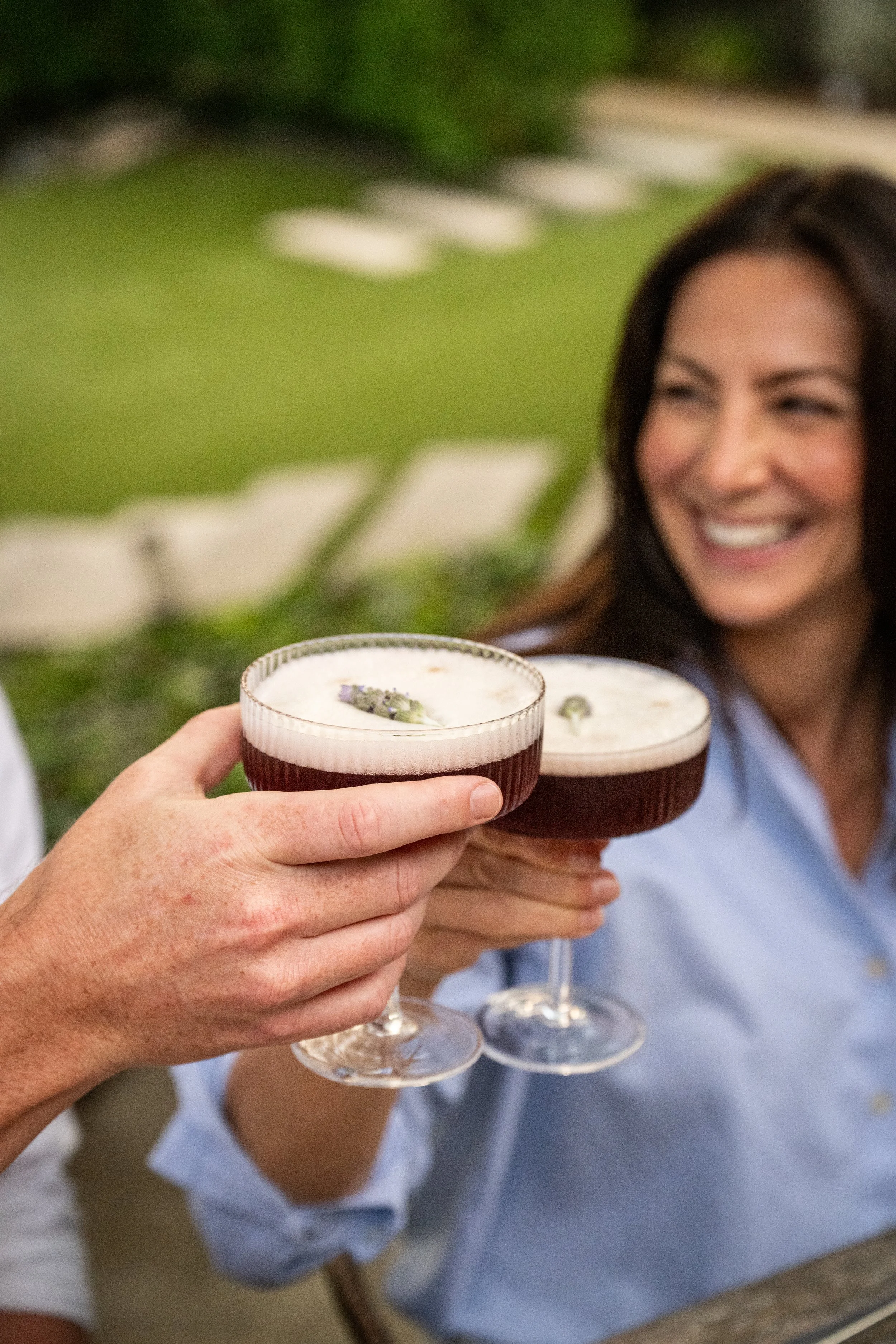 Two people clinking glasses of dark red wine or cocktail outdoors, with a woman smiling in the background.