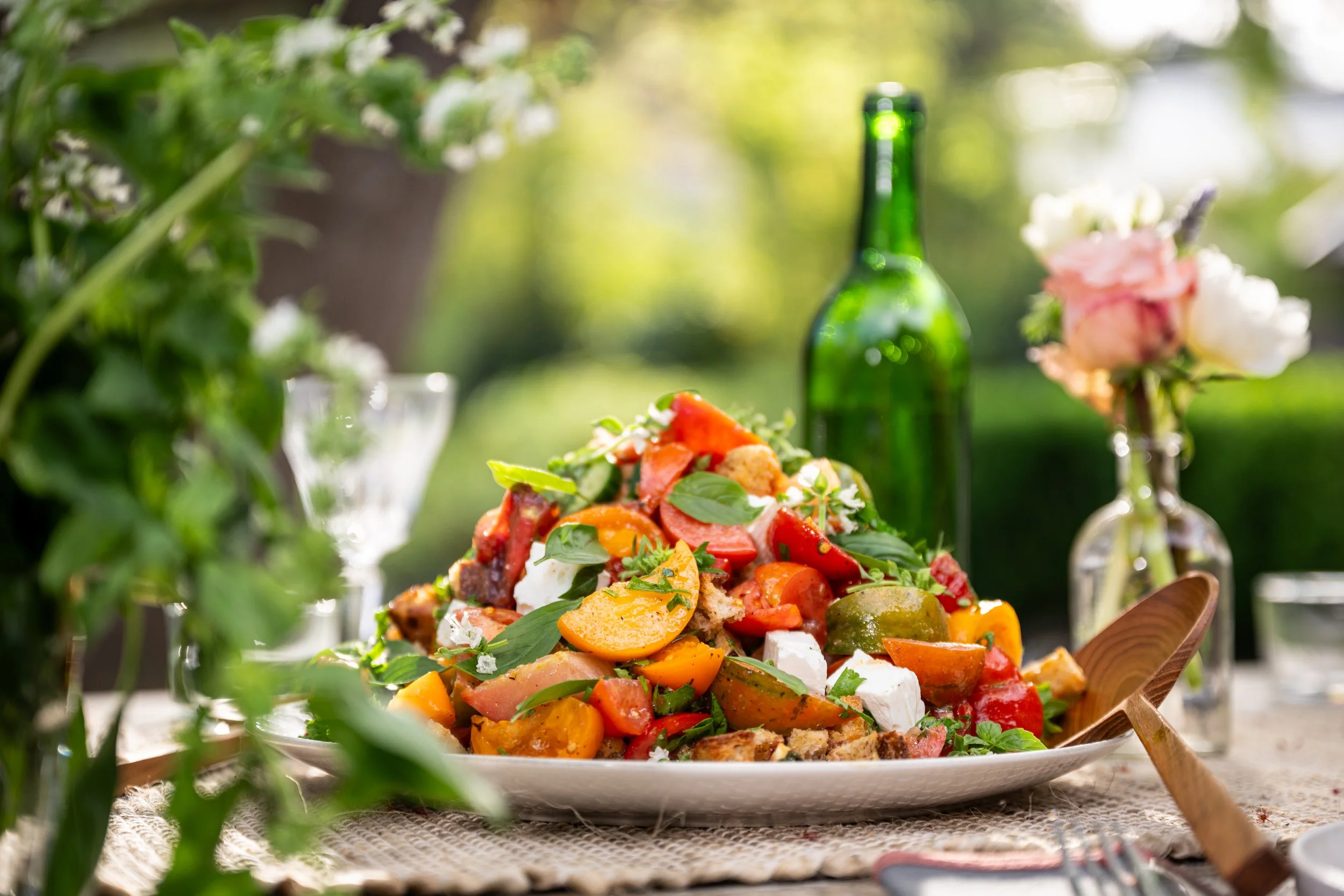 Fresh vegetable salad with cherry tomatoes, cucumbers, feta cheese, and basil leaves on a white plate, set outdoors with a green bottle and a flowered vase in background.