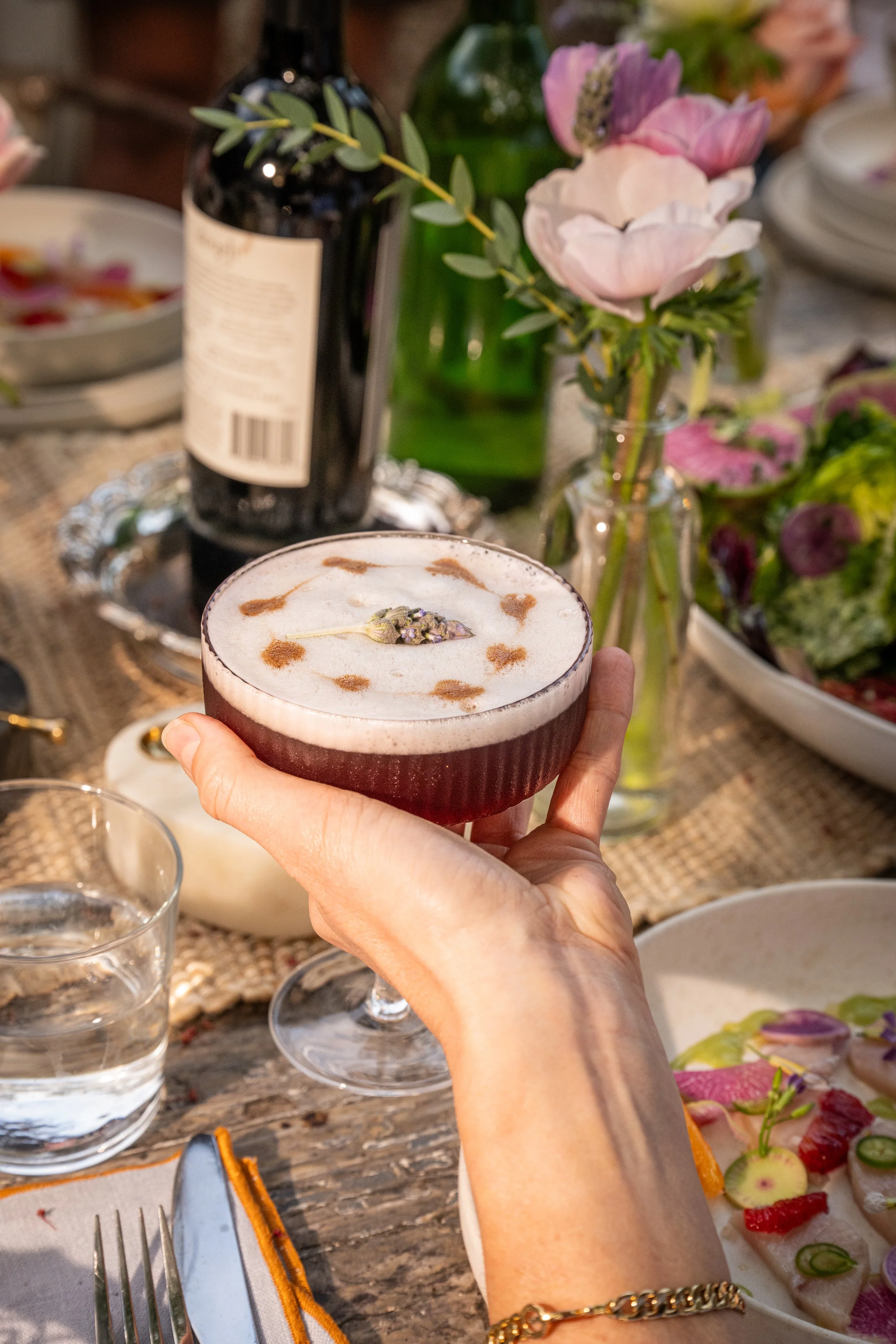 A person holding a cocktail glass with frothy foam and lavender garnish at a dinner table with flowers, salads, bottled drinks, and glass of water.
