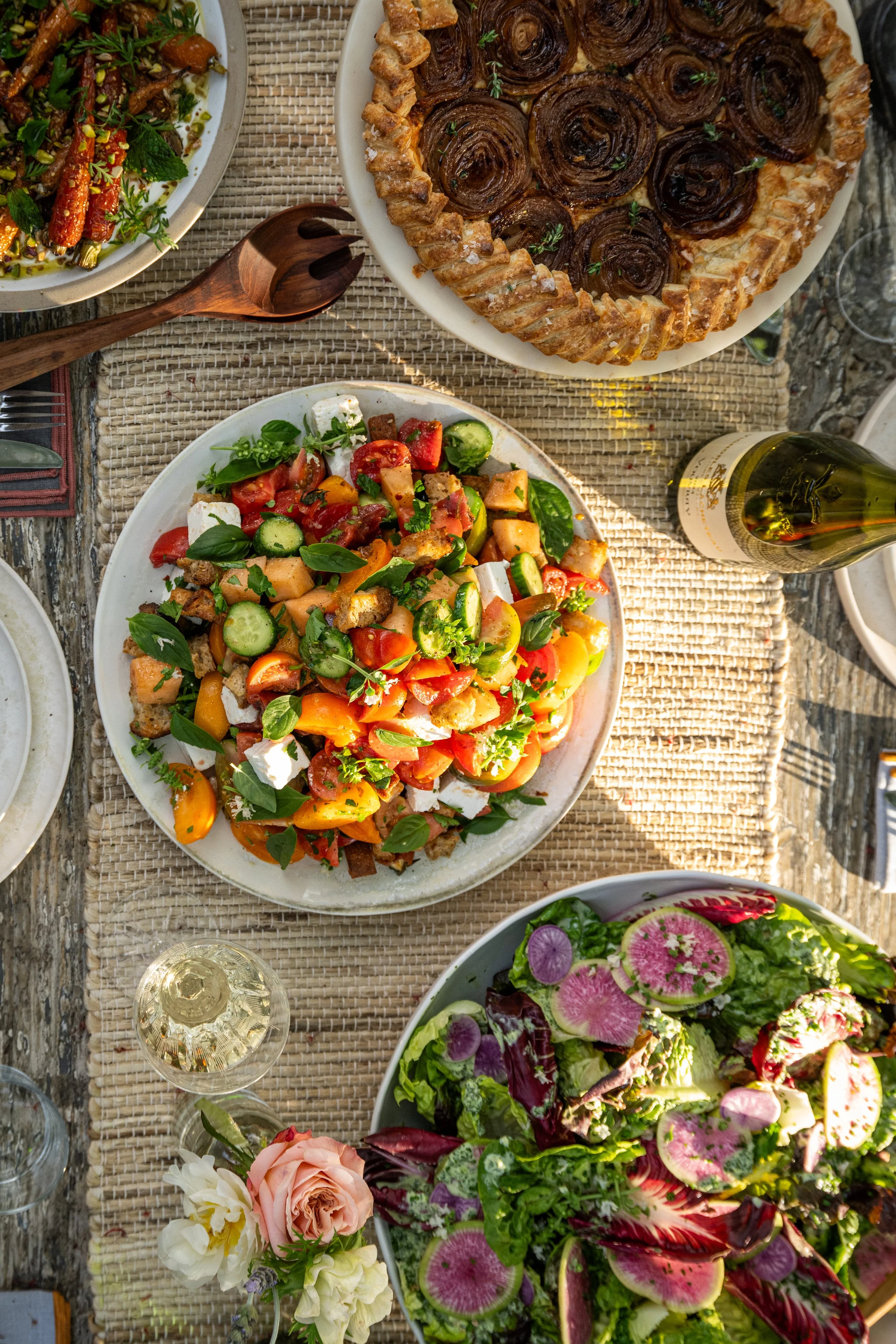 A table set with various salads and a pie, featuring a fresh vegetable salad with tomatoes, cucumbers, basil, and cheese, a mixed greens salad with watermelon radishes, and a caramelized onion tart.