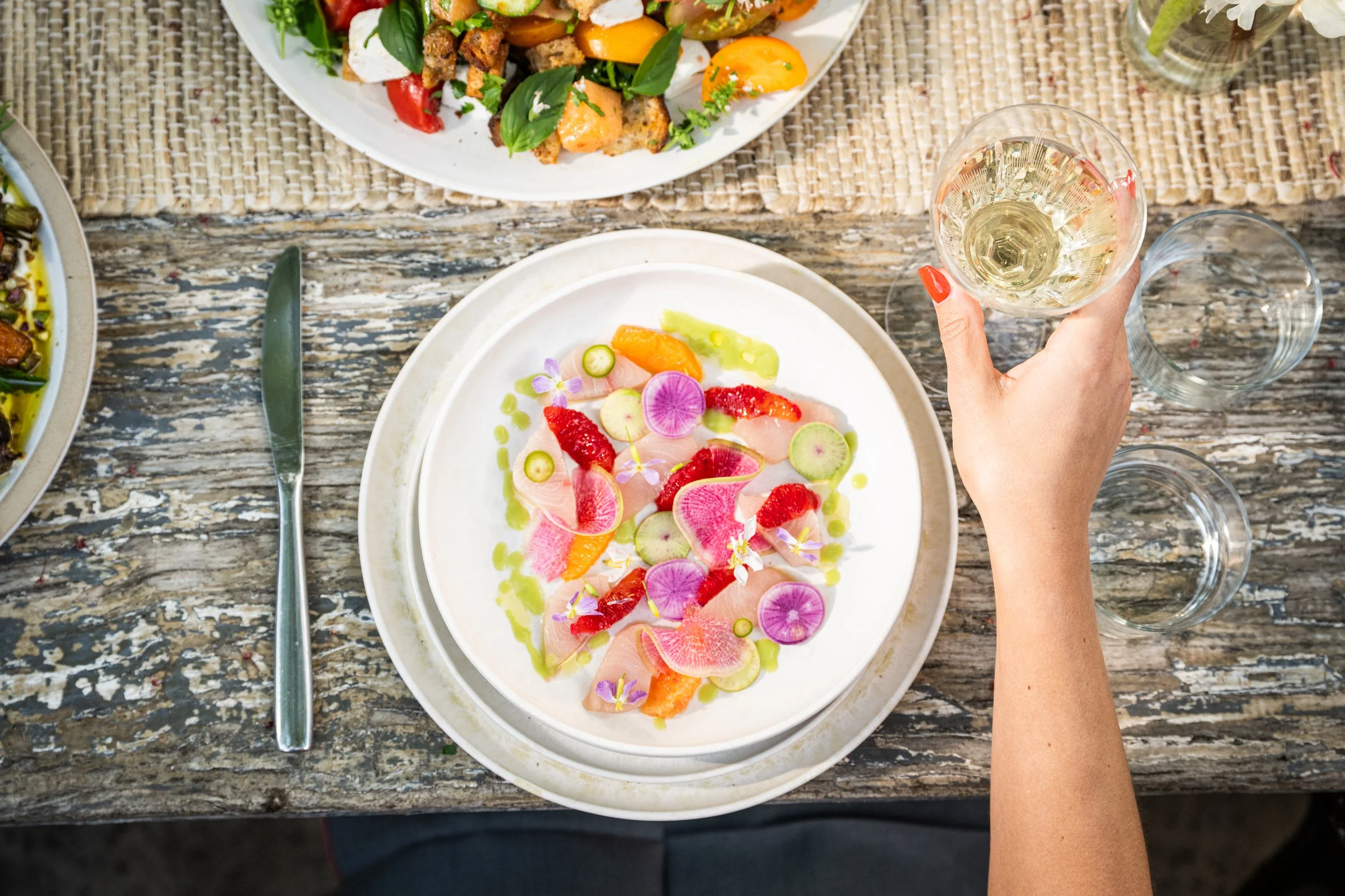 A person holding a glass of white wine over a rustic wooden table with a white plate of colorful sliced vegetables and edible flowers, with other plates of food visible.