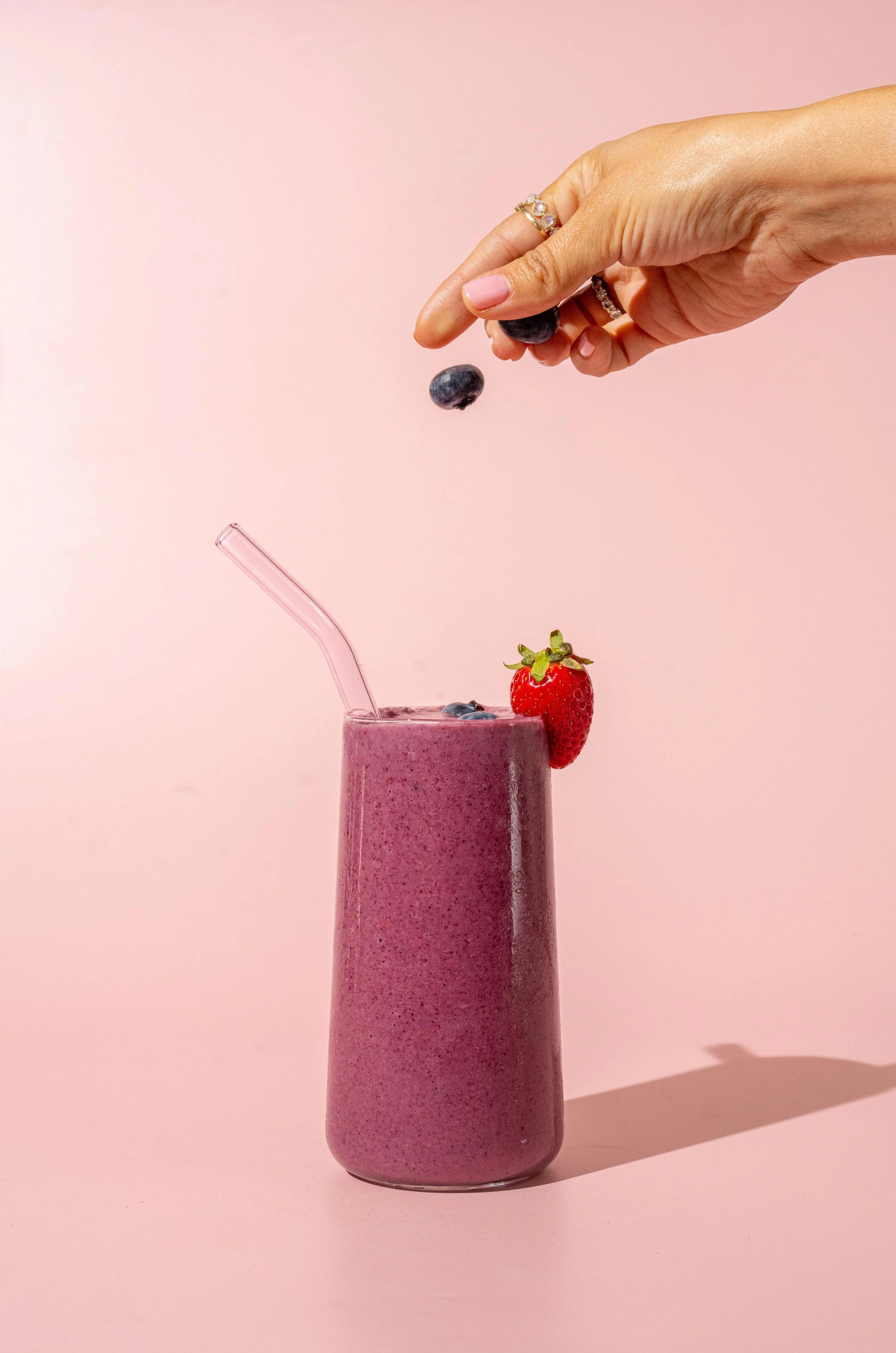 A hand adding blueberries to a strawberry smoothie in a tall glass with a pink background.