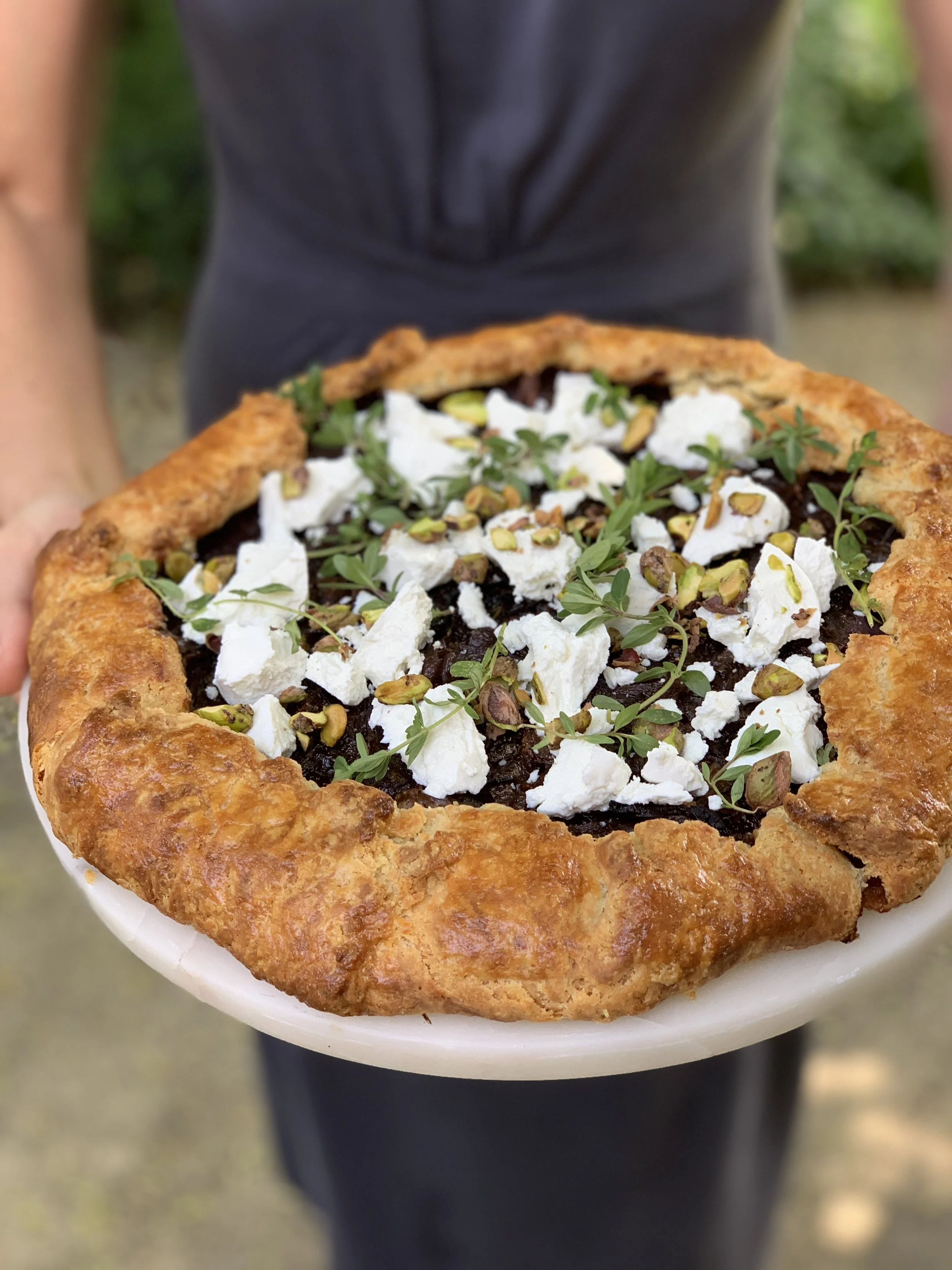 Close-up of a person holding a savory pie topped with crumbled cheese, chopped herbs, chopped nuts, and a dark filling, with a blurred outdoor background.