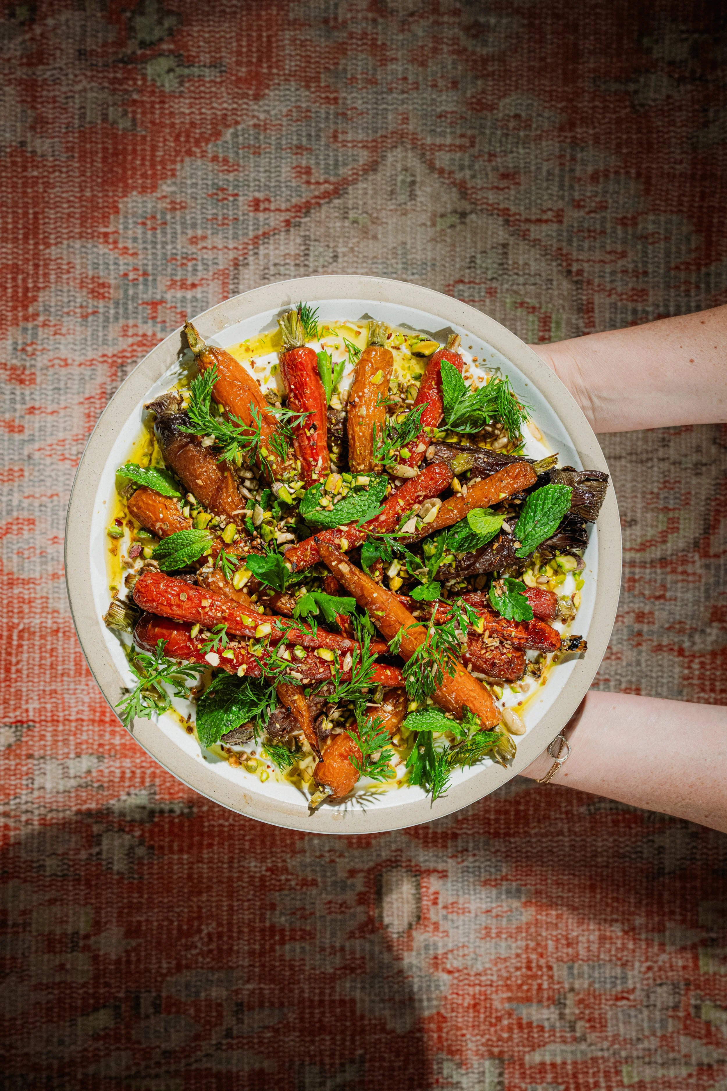 A person holding a round white dish filled with roasted carrots, garnished with fresh herbs, chopped nuts, and a drizzle of oil, on a patterned rug.