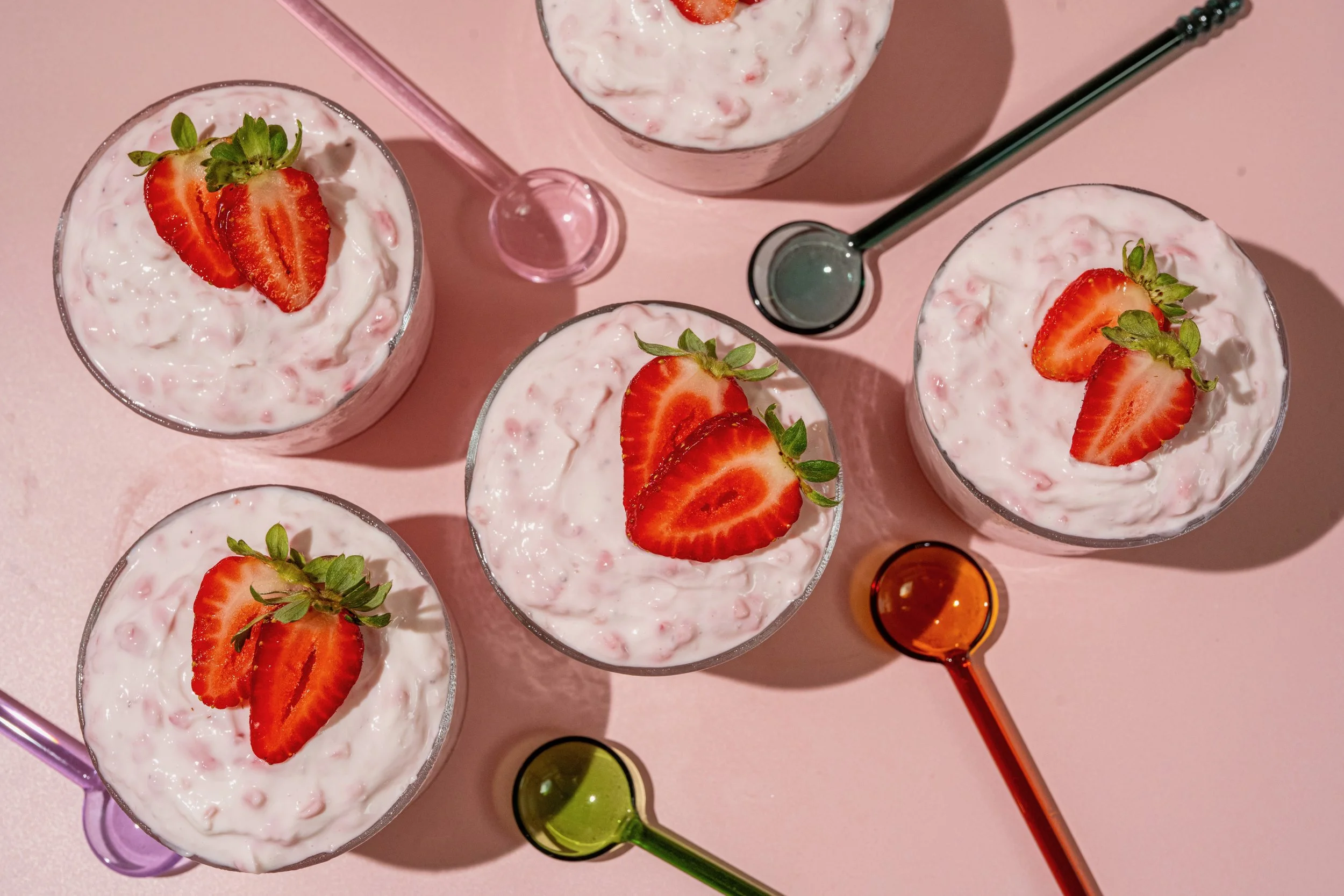 Five glasses of strawberry dessert topped with sliced strawberries and mint on a pink surface, with colorful glass straws and small bottles nearby.