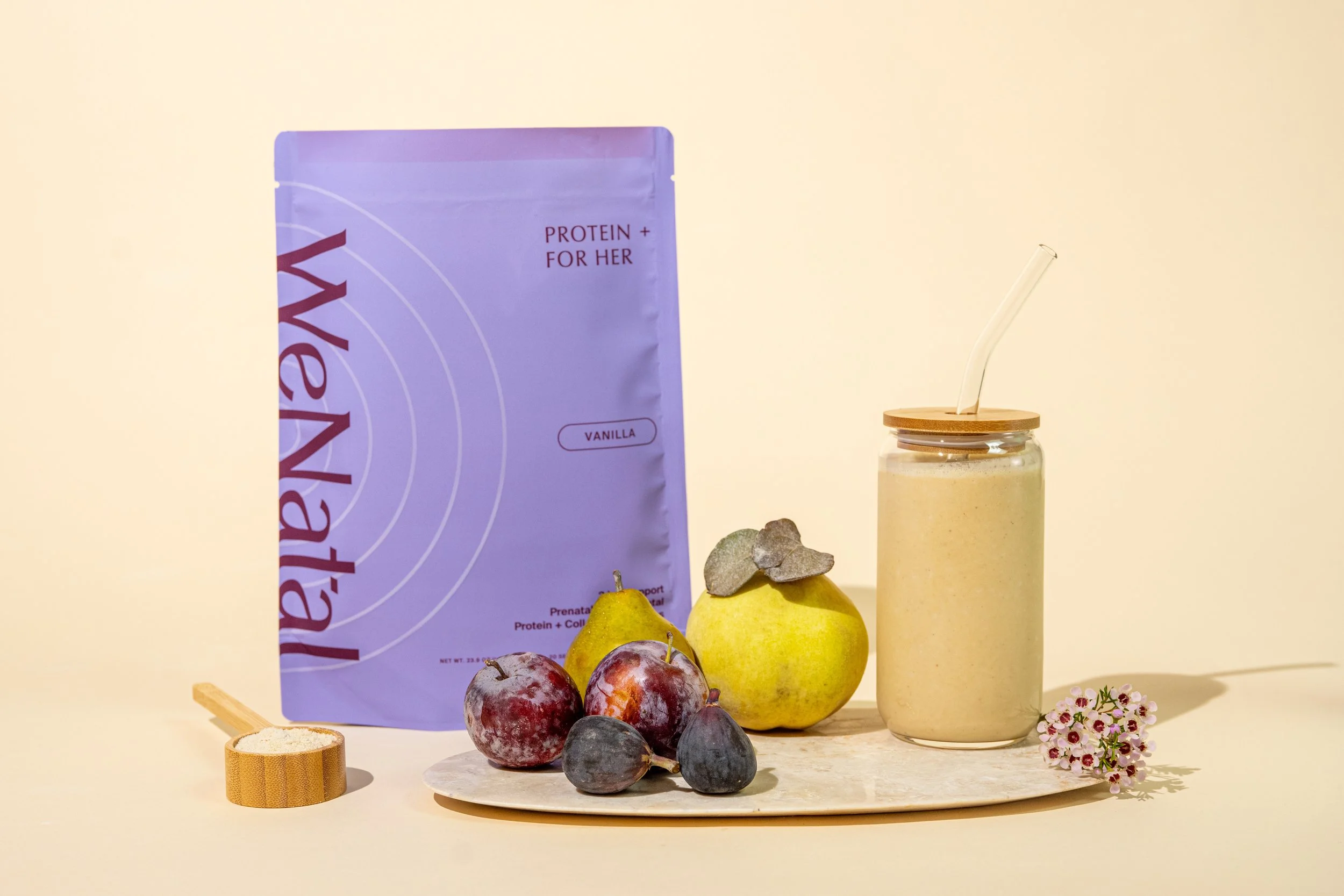 A purple package labeled 'WeNady' with 'protein + for her', a jar with a straw, assorted fruits including apples, pears, and plums, a small scoop of powder, and a small bouquet of flowers on a round beige surface against a neutral background.