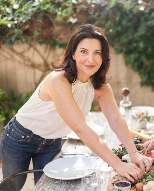 A woman with shoulder-length dark hair, wearing a sleeveless cream top and blue jeans, is preparing food outdoors at a table with various ingredients and utensils, smiling at the camera.
