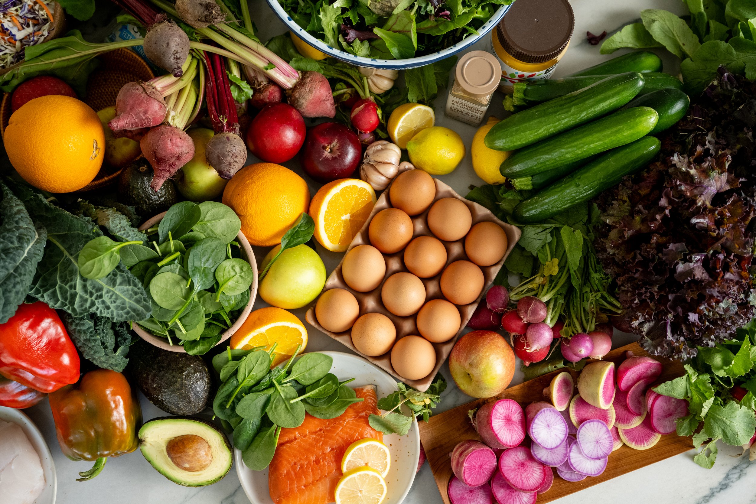 A variety of fresh produce including oranges, lemons, radishes, cucumbers, eggs, leafy greens, an avocado, a salmon fillet, radishes, and other vegetables and herbs arranged on a table.