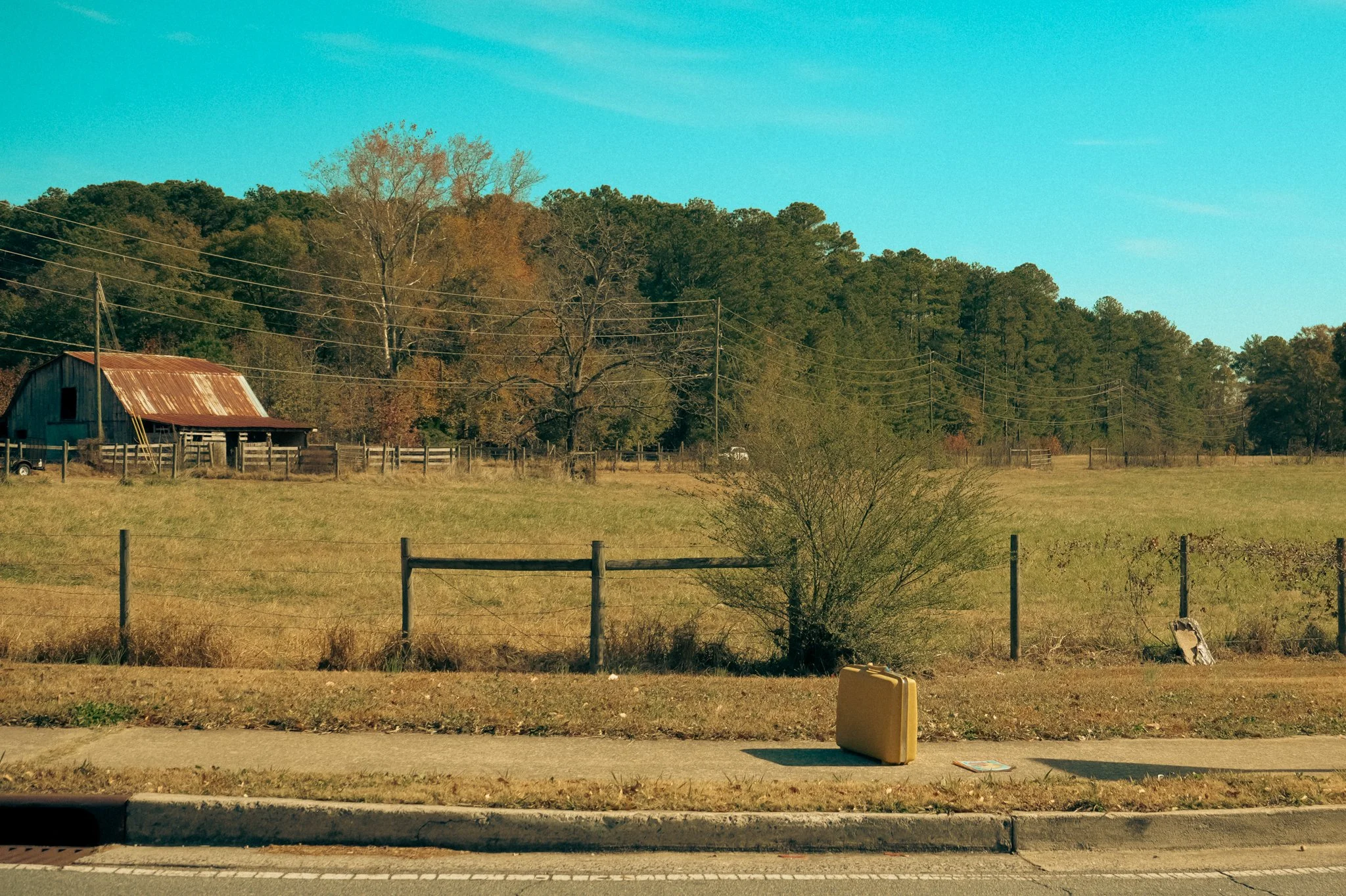 A rural scene with a grassy field, a rusty barn, trees in autumn foliage, power lines, a fence, and a sidewalk with a yellow utility box and a paper on the ground under a blue sky.