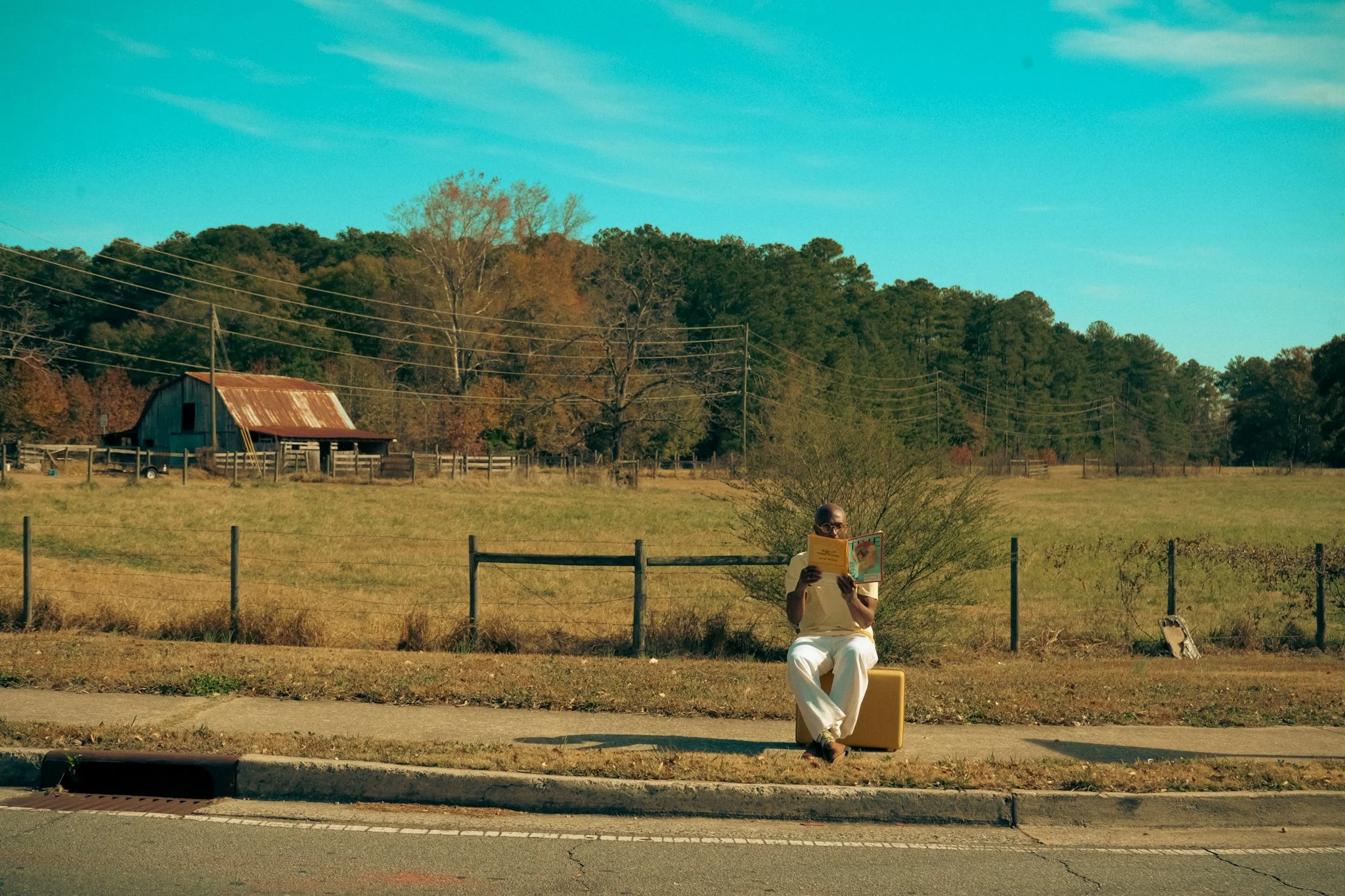 Person sitting on a yellow suitcase, reading a book, in front of a rural landscape with trees, a weathered barn, and power lines under a clear blue sky.