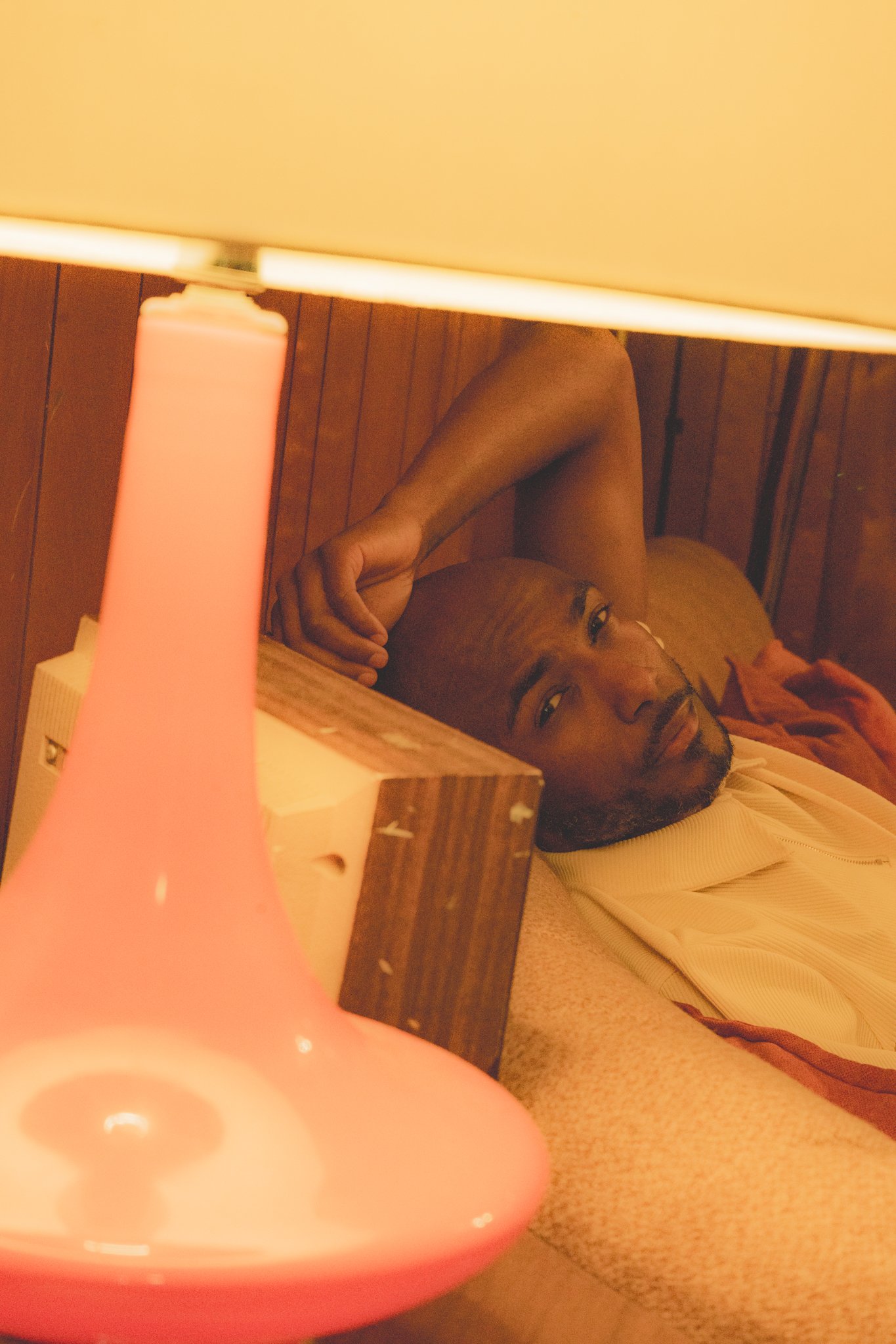 A man with glasses and a beard lying on the floor, partially under a table, looking at the camera. The image is taken from a low angle with a pink lamp in the foreground.