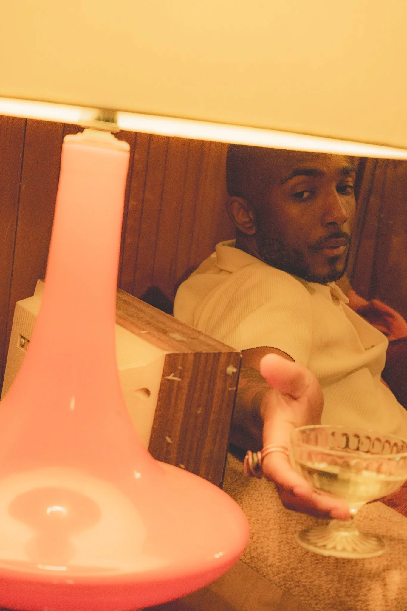 A person with dark hair and facial hair is sitting indoors with a wood-paneled wall behind them. They are wearing a light-colored shirt and looking at the camera. In the foreground, there is a pink lamp and a glass of beer held by someone else.