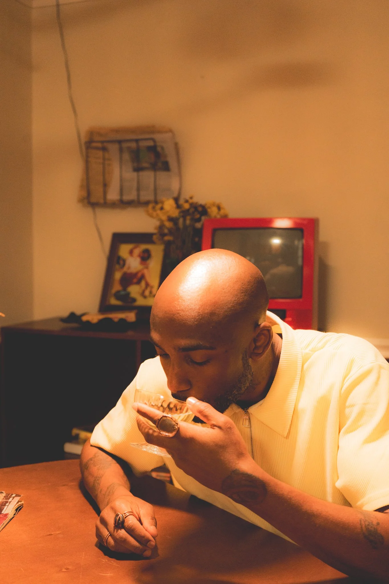 A man with a shaved head and tattoos on his arms, wearing a yellow shirt, is sitting at a wooden table, drinking from a glass. The background includes a framed picture, a TV, a decorative shelf with flowers, and wall decorations.