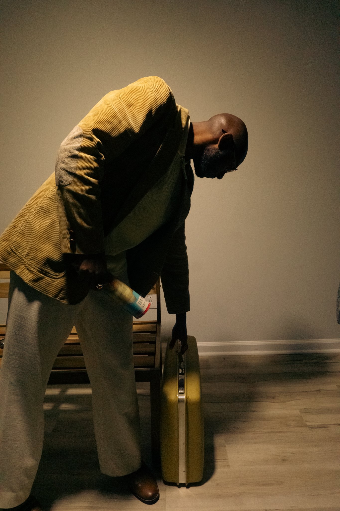 A person with dark skin, wearing glasses, a beige jacket, and gray pants, is holding a yellow suitcase in front of a mannequin. The person is looking down and appears to be in an indoor setting with gray walls and wooden flooring.