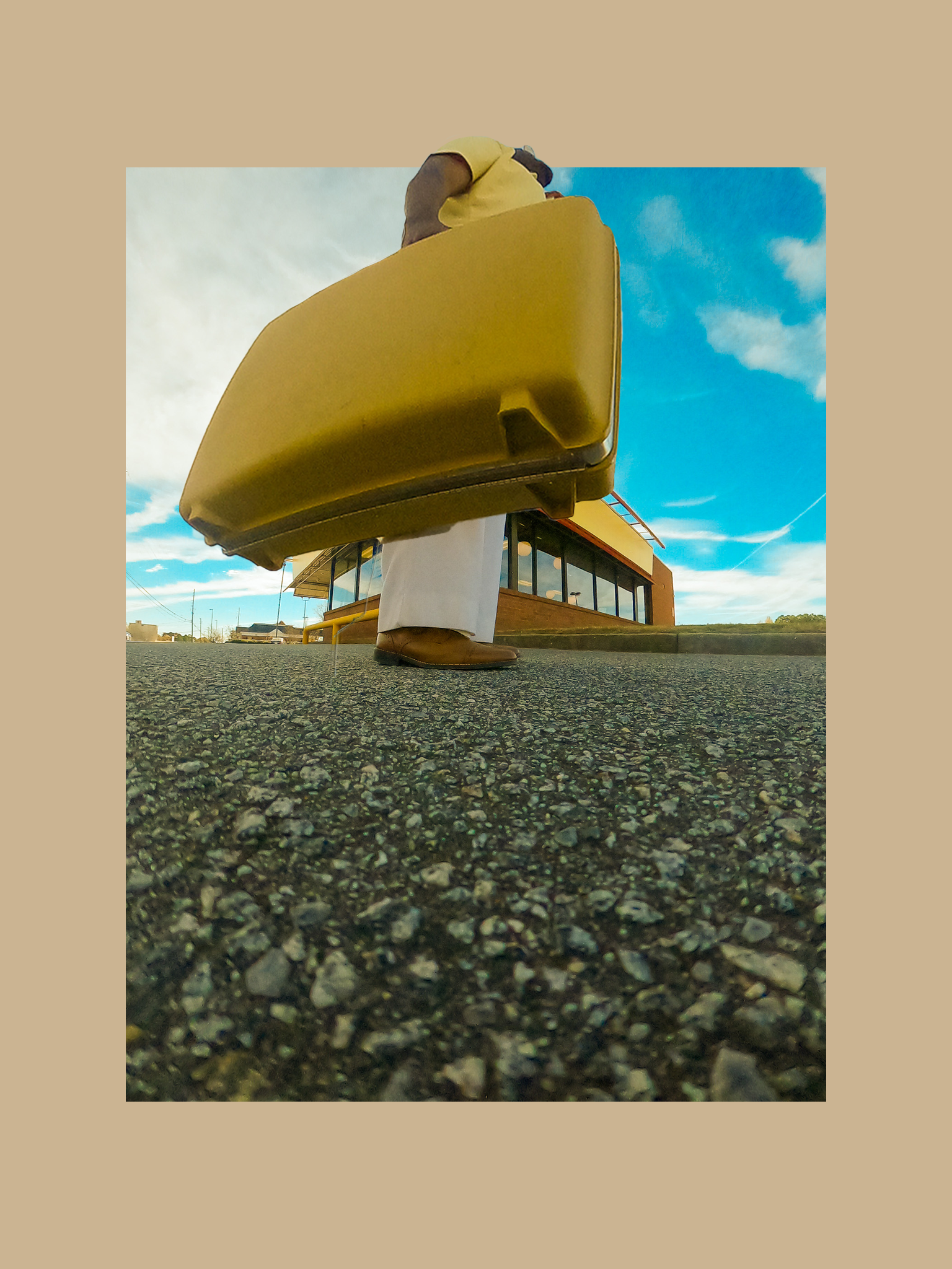 Low-angle photo of a woman with dark hair wearing a yellow shirt and white pants, standing on a pebbled surface with a yellow building behind her, under a partly cloudy sky.