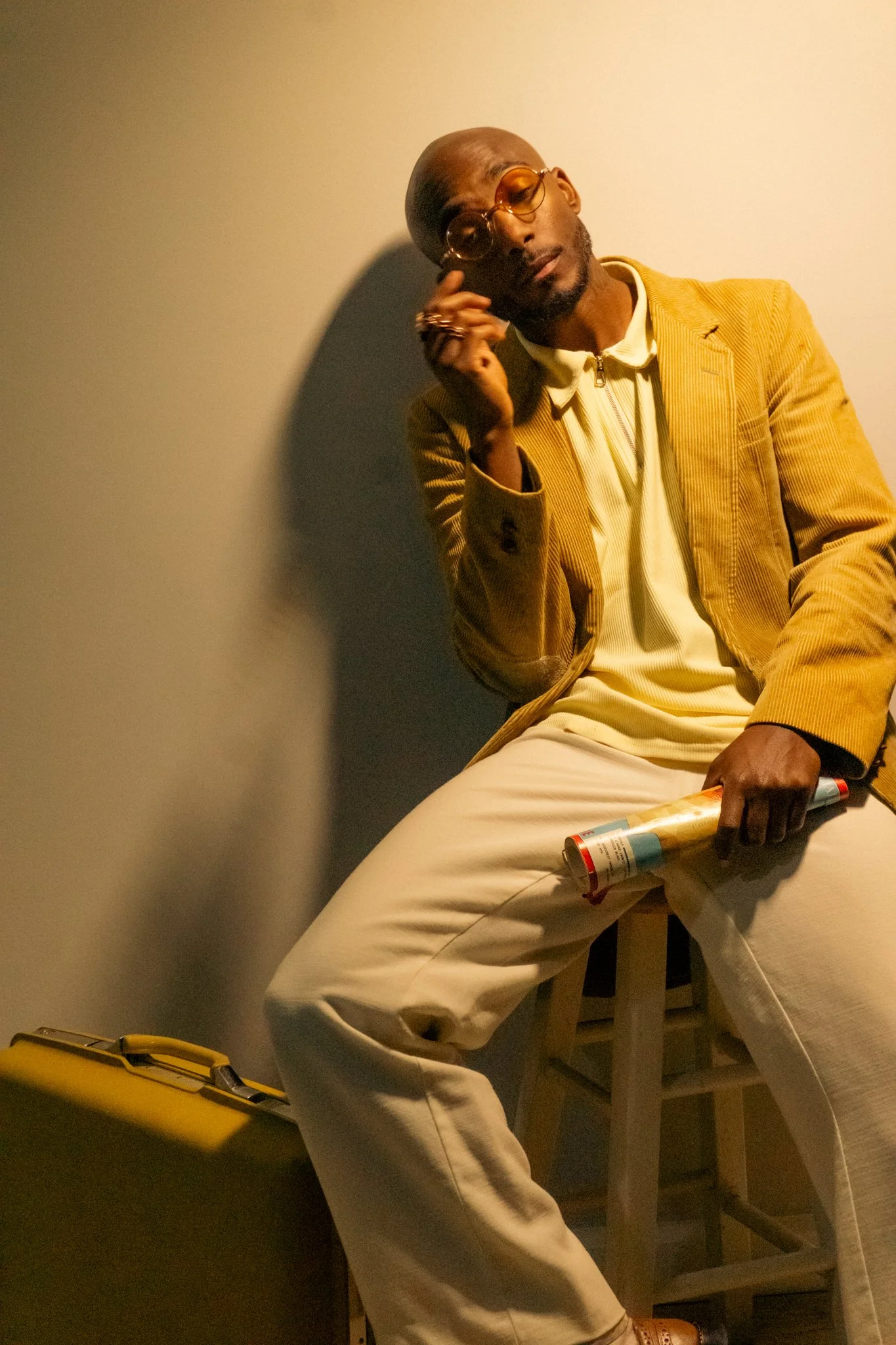 A man with a bald head, glasses, and a beard, dressed in a yellow corduroy jacket, cream shirt, and white pants, sitting on a stool against a light-colored wall. He is holding a spray can in his right hand and appears to be touching his head with his left hand, with a thoughtful expression.