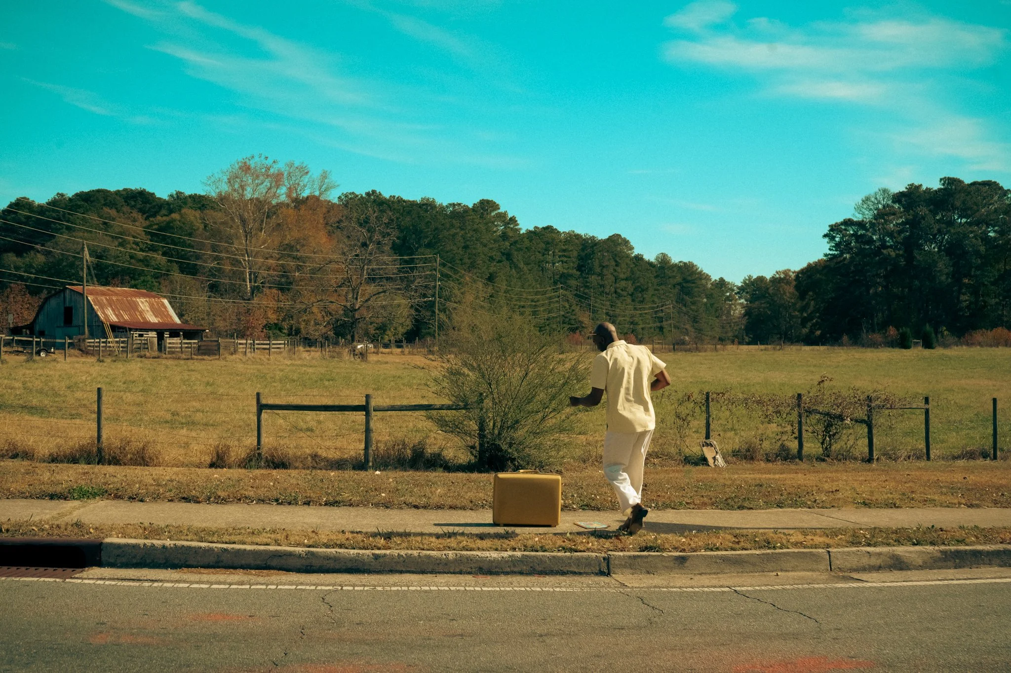 A person in white clothing planting a small tree in a grassy field, with a rustic barn and power lines in the background under a blue sky.