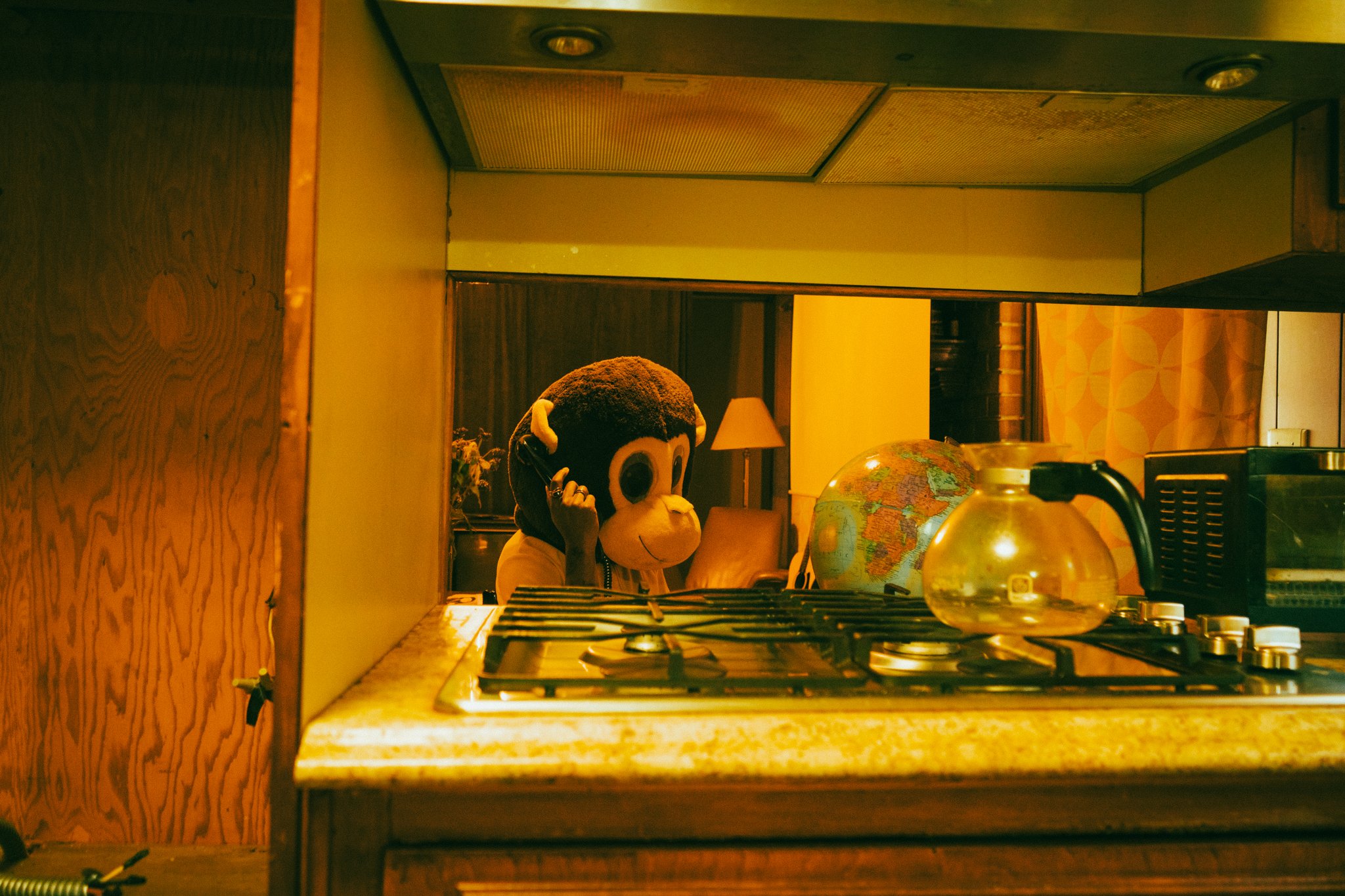 A person wearing a monkey mascot head mask talking on the phone in a room with a globe, lamp, and kitchen items on counter, seen through a window in a kitchen.