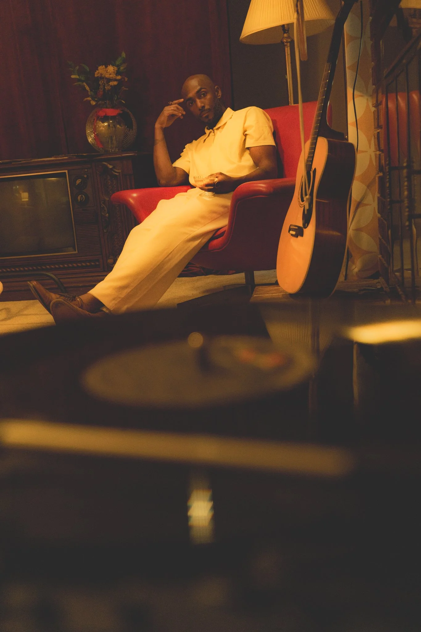 A man sitting on a red chair in a dimly lit room, with a guitar resting against the chair, a vintage television, a lamp, and a vase with flowers on a cabinet in the background.