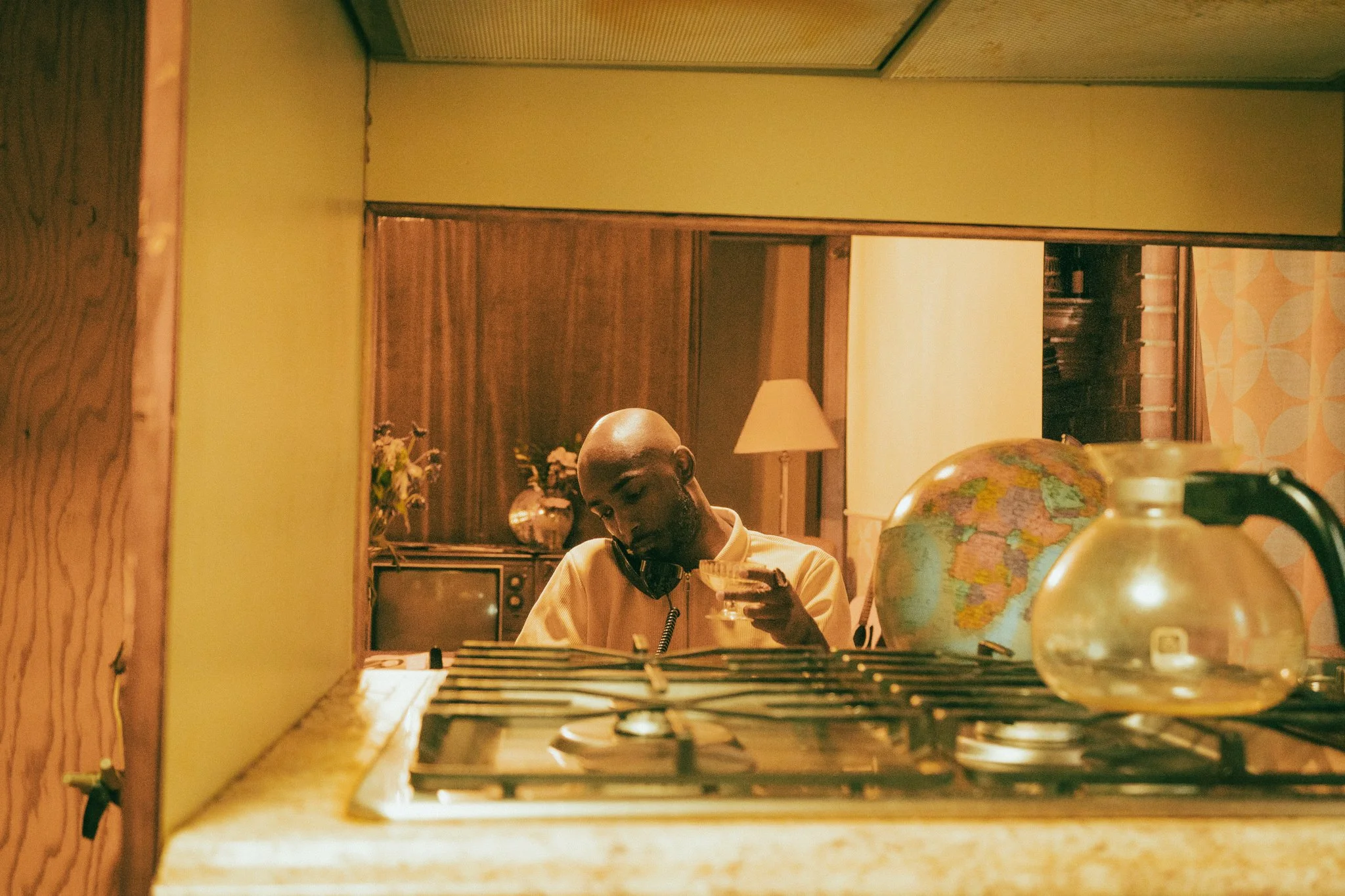 A man sitting at a table, wearing a yellow shirt and headphones, looking at his phone. There is a globe on the table, a lamp in the background, and a vintage television set.
