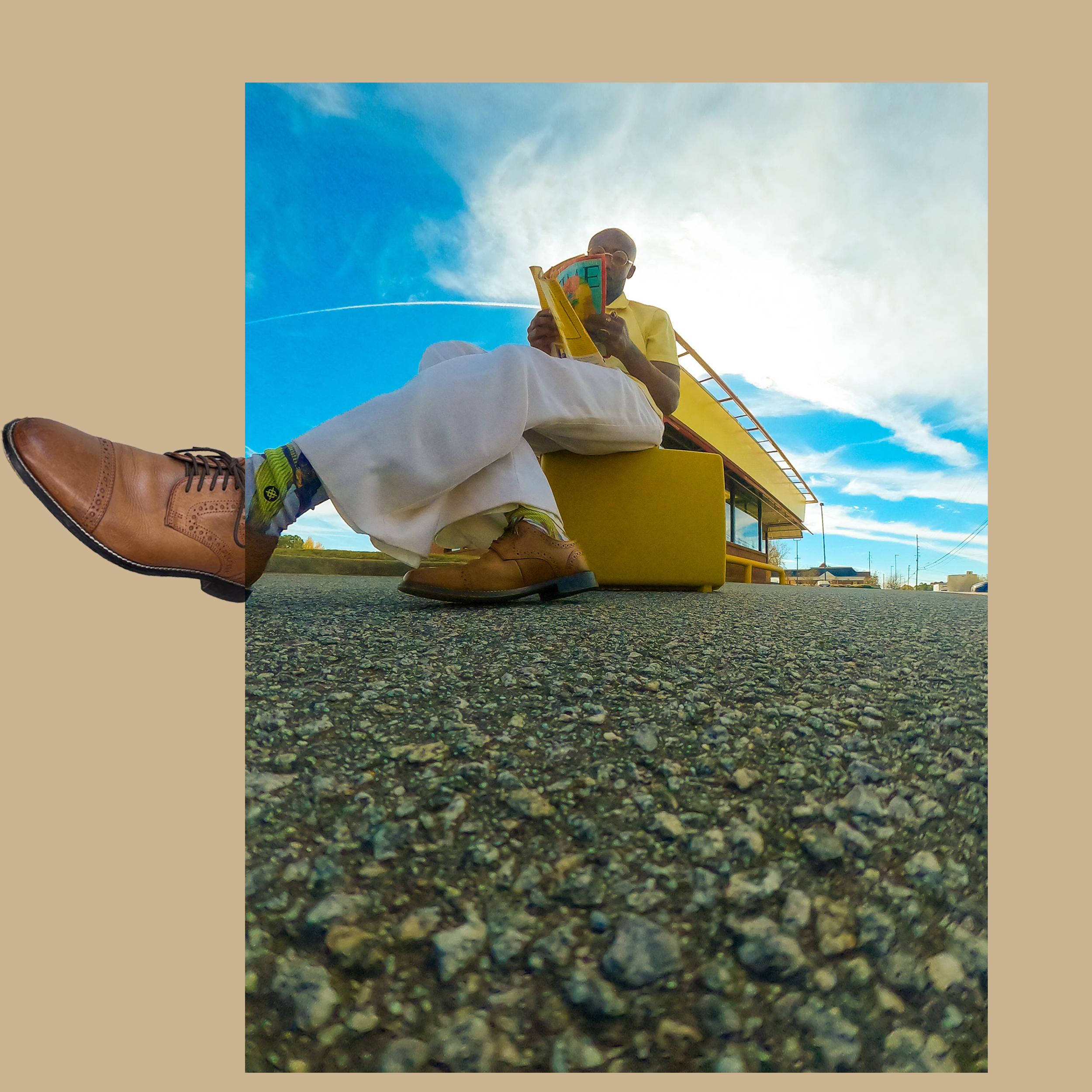 A person's foot in a brown dress shoe with laces standing on a gravel surface, with a man sitting cross-legged reading a colorful book next to a yellow structure under a partly cloudy sky.
